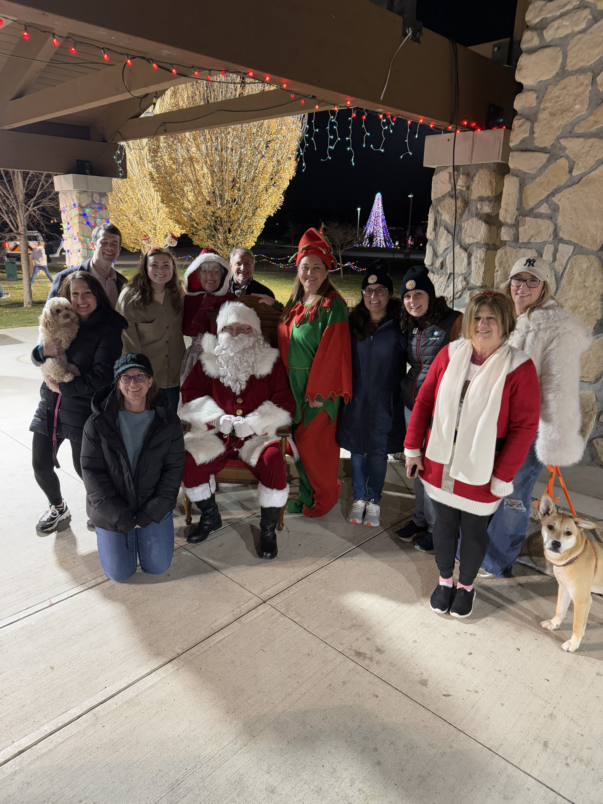 Group of people dressed in holiday costumes and winter clothing, including Santa Claus, Mrs. Claus, an elf, and others, gathered outside at night with Christmas lights and a decorated tree in the background, along with dogs.