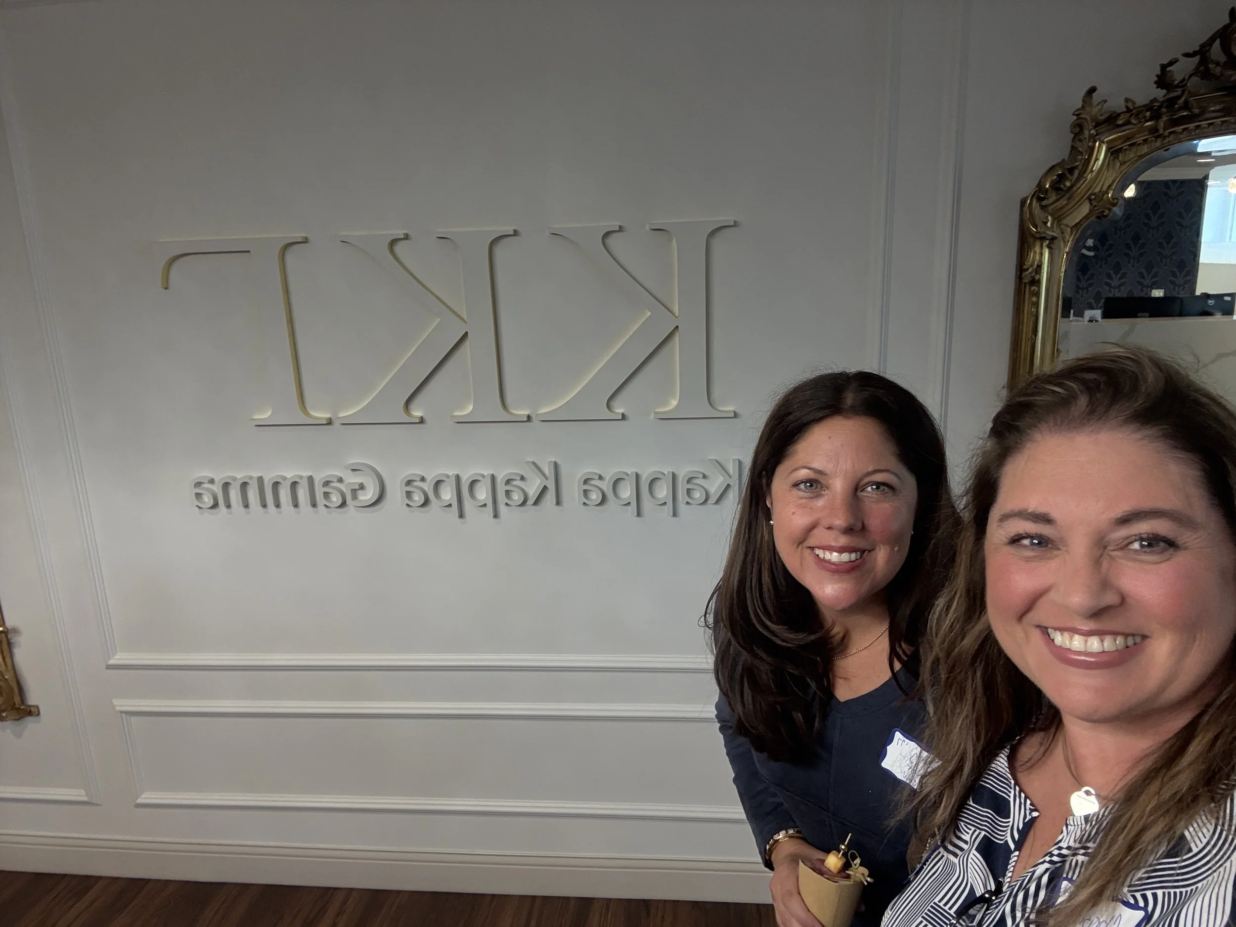 Two smiling women standing in front of a wall with the words "KAPPA KAPPA GAMMA" in raised letters, next to a large ornate mirror, in a room with white walls and wooden flooring.