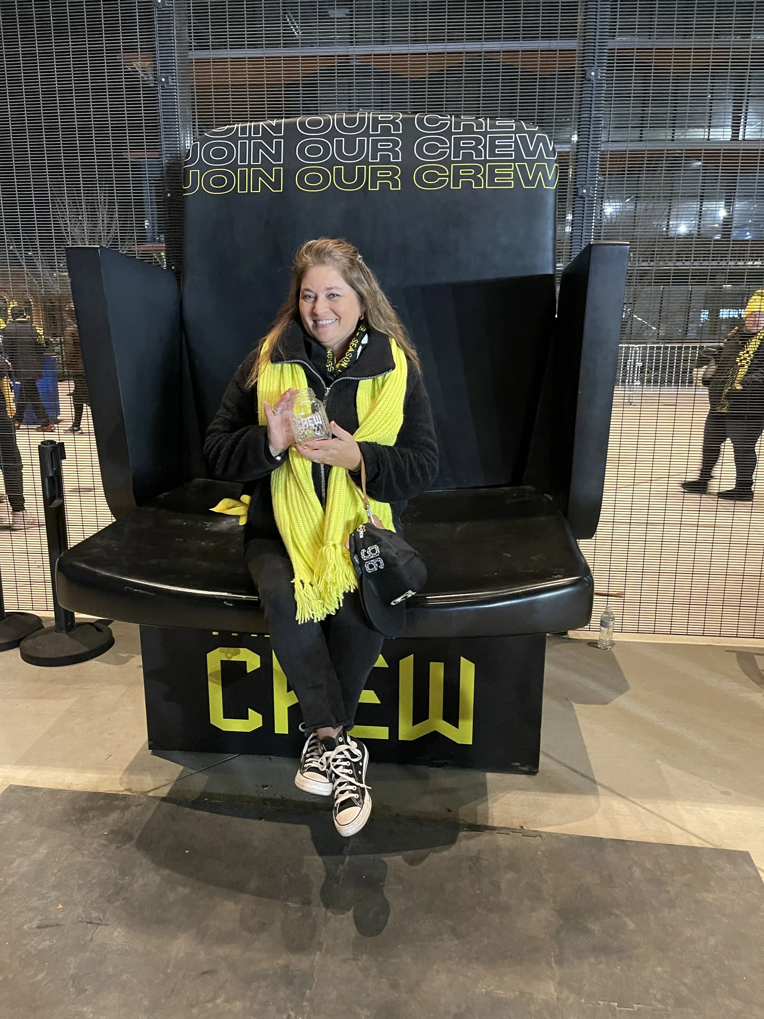 A woman sitting on a black seat with yellow 'Crew' text, in front of a large black display with 'Join Our Crew' written repeatedly, holding a clear cup with a drink, indoors with a metal wire fence background and others in the distance.
