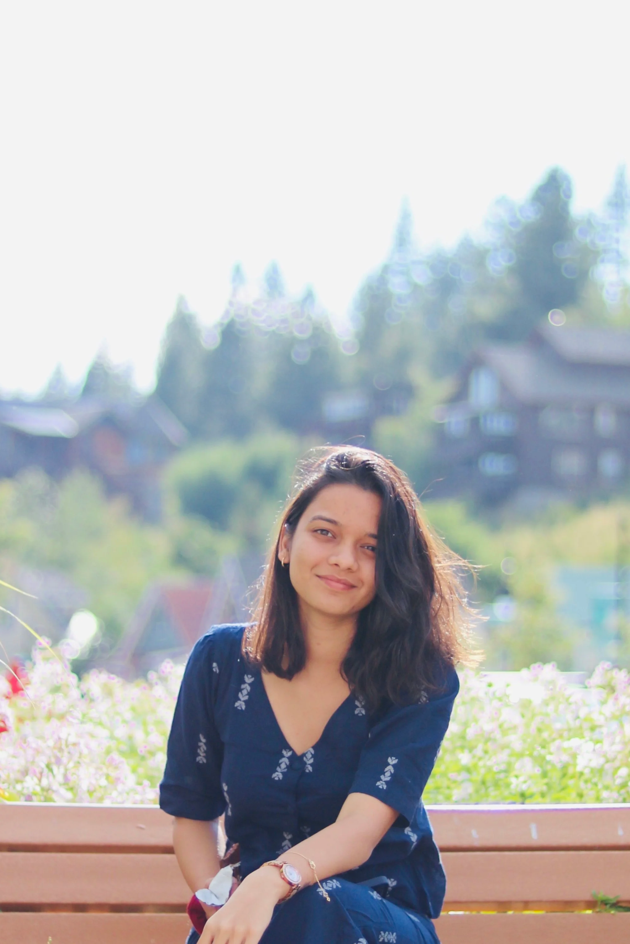 A woman with dark brown hair and a light smile sitting on a wooden bench outdoors, with out-of-focus houses and trees in the background illuminated by sunlight.