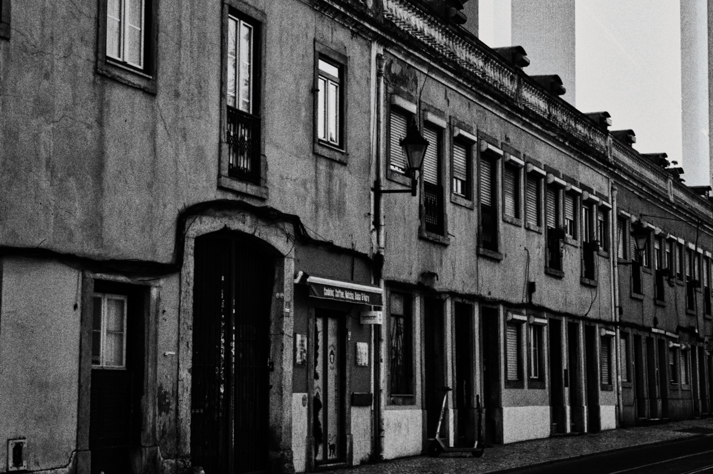 Rua com edifícios antigos de fachada envelhecida, janelas com grades e uma porta de entrada de arco. Uma lâmpada de rua e uma placa de informação ao lado da entrada. A imagem é em preto e branco.