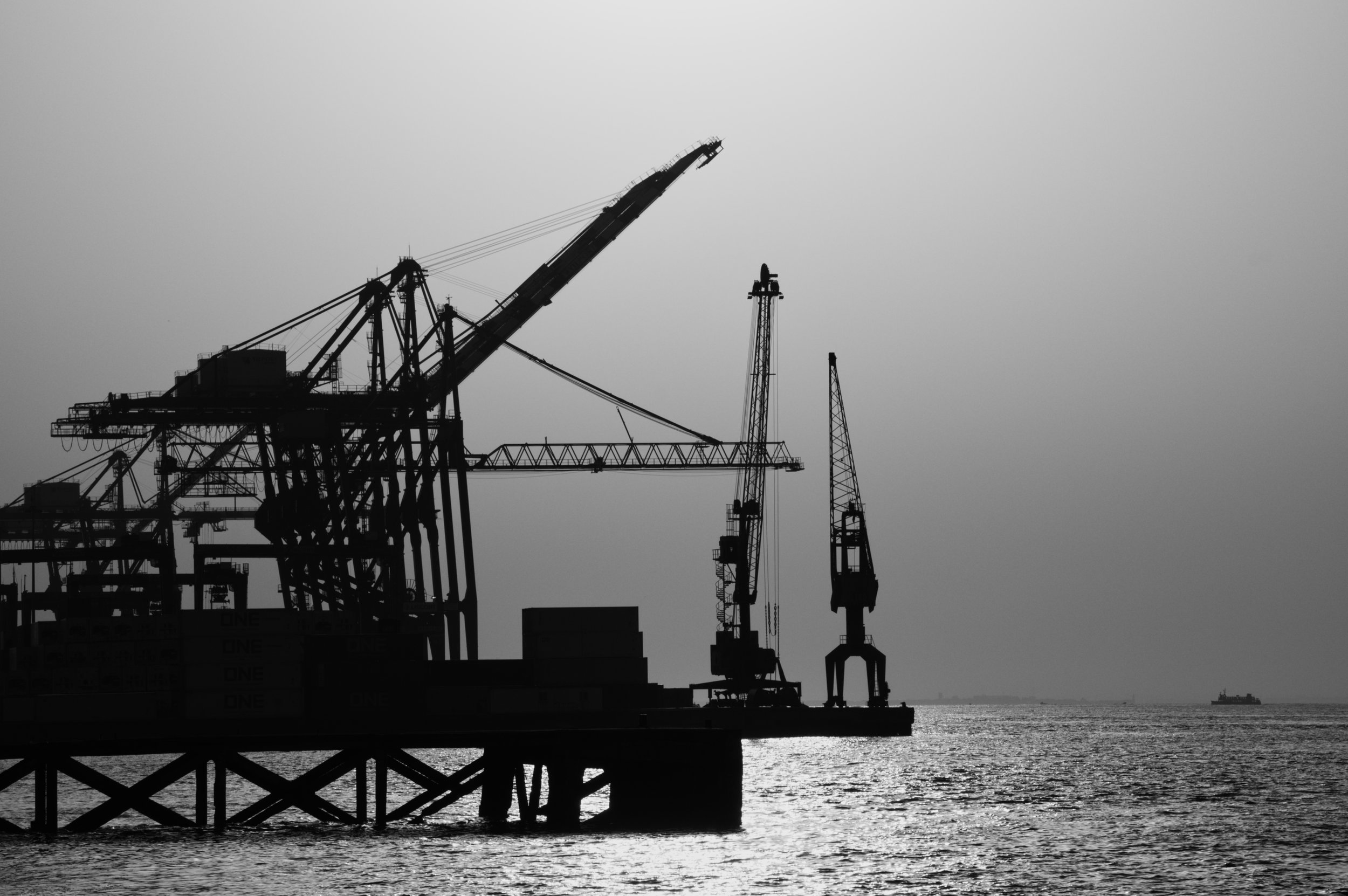 Silhueta de plataformas de petróleo e guindastes no mar ao entardecer, com um navio ao fundo no horizonte.
