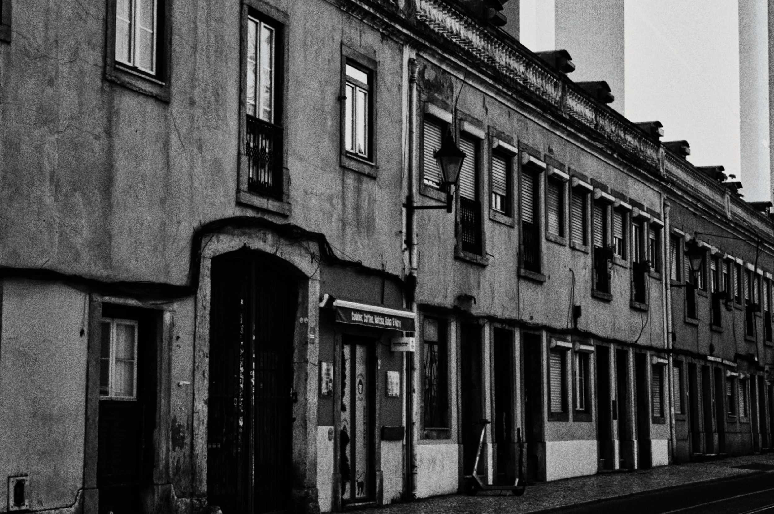 Rua de parede antiga com várias janelas e portas, algumas com grades e cortinas. Há uma placa de rua e luminárias na parede. A cena é em preto e branco.