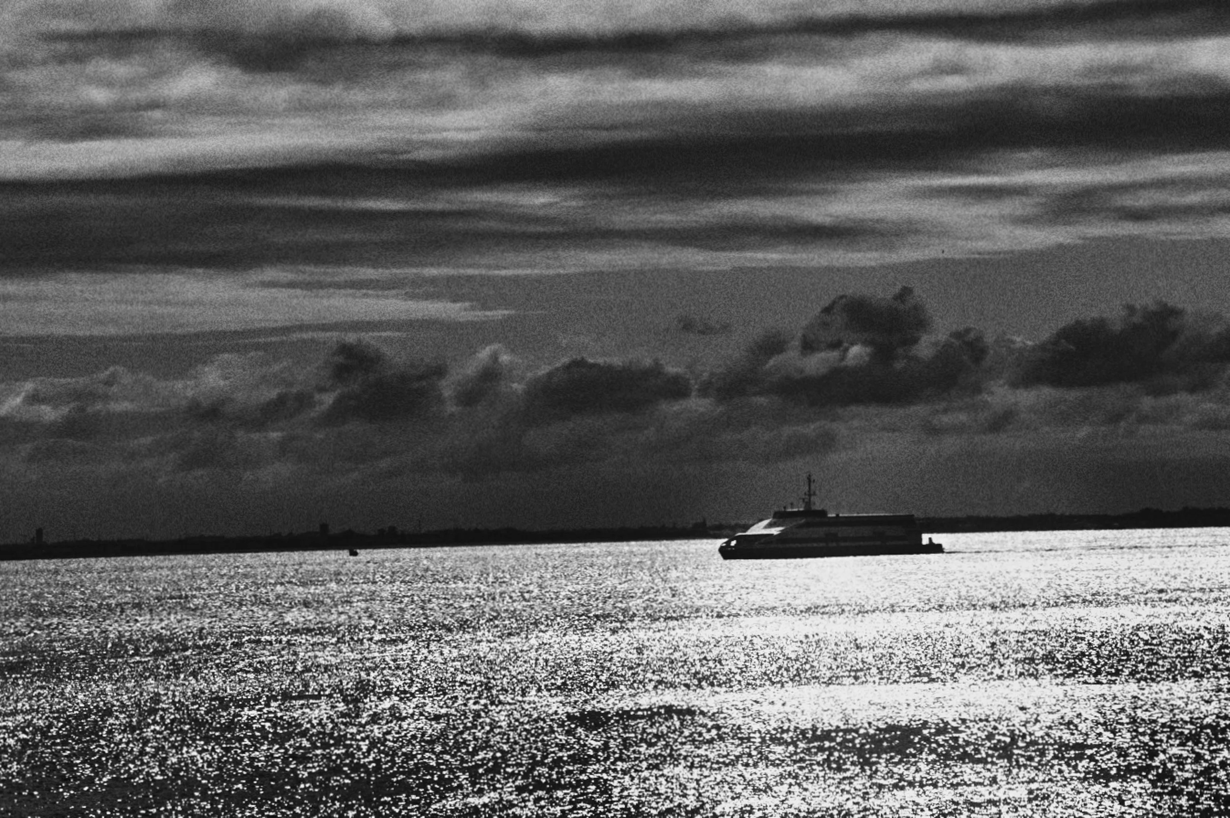 Imagem em preto e branco de um mar ou rio com um barco ao longe e nuvens carregadas no céu.
