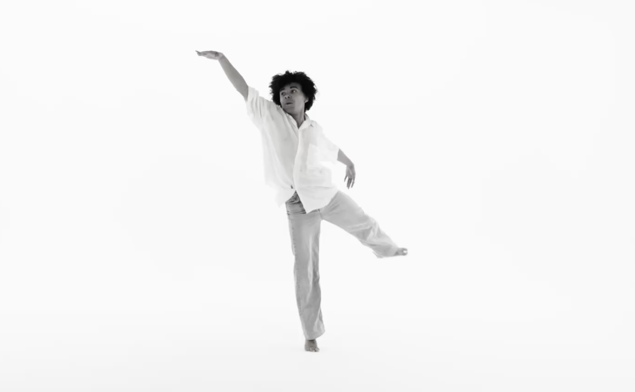 Une personne danse en position équilibrée, portant des vêtements blancs sur fond blanc. L'image est en noir et blanc.