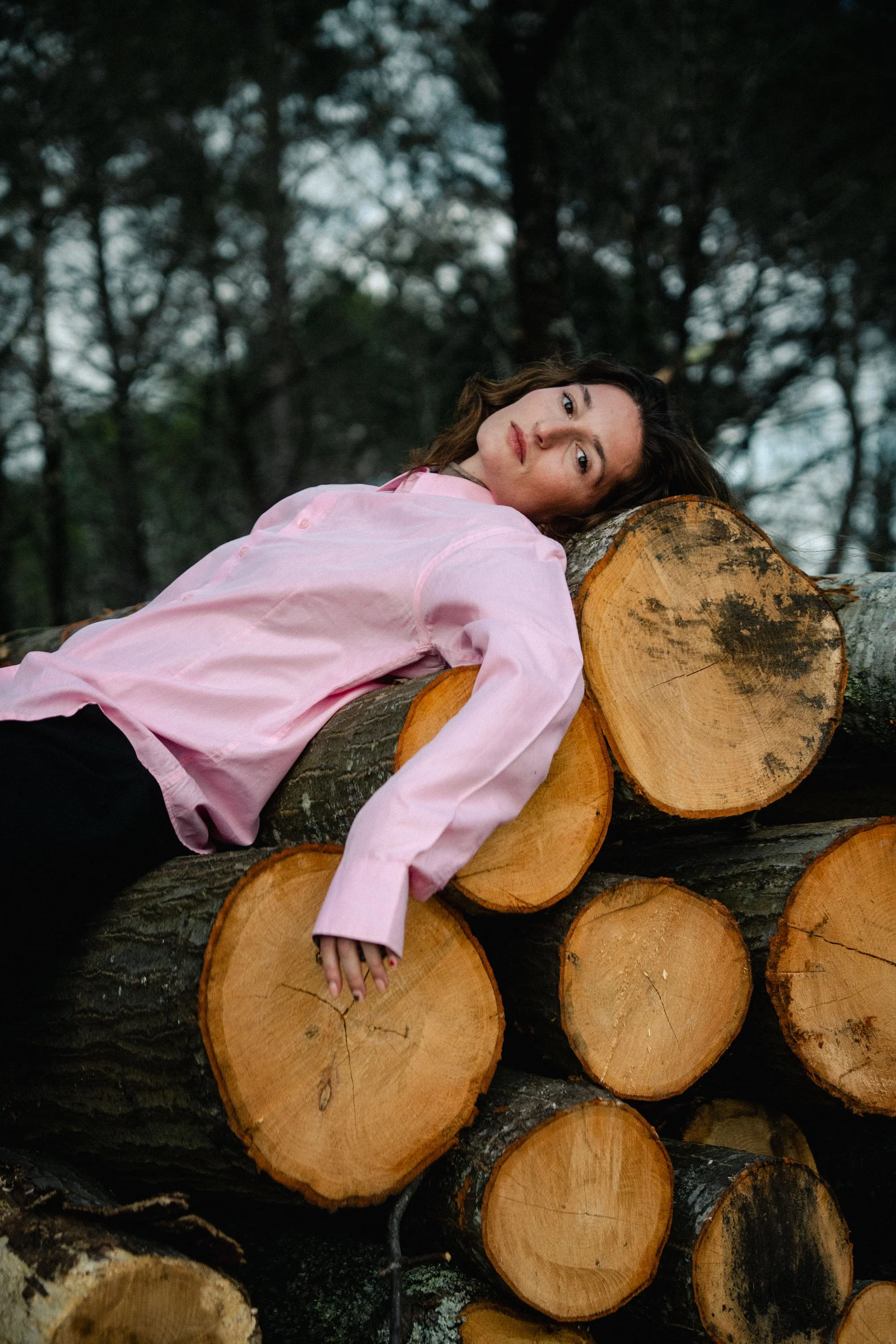 Une femme repose sa tête sur une bûche d'arbre coupée dans une forêt, portant une chemise rose clair, avec des arbres en arrière-plan, ambiance naturelle et calme.