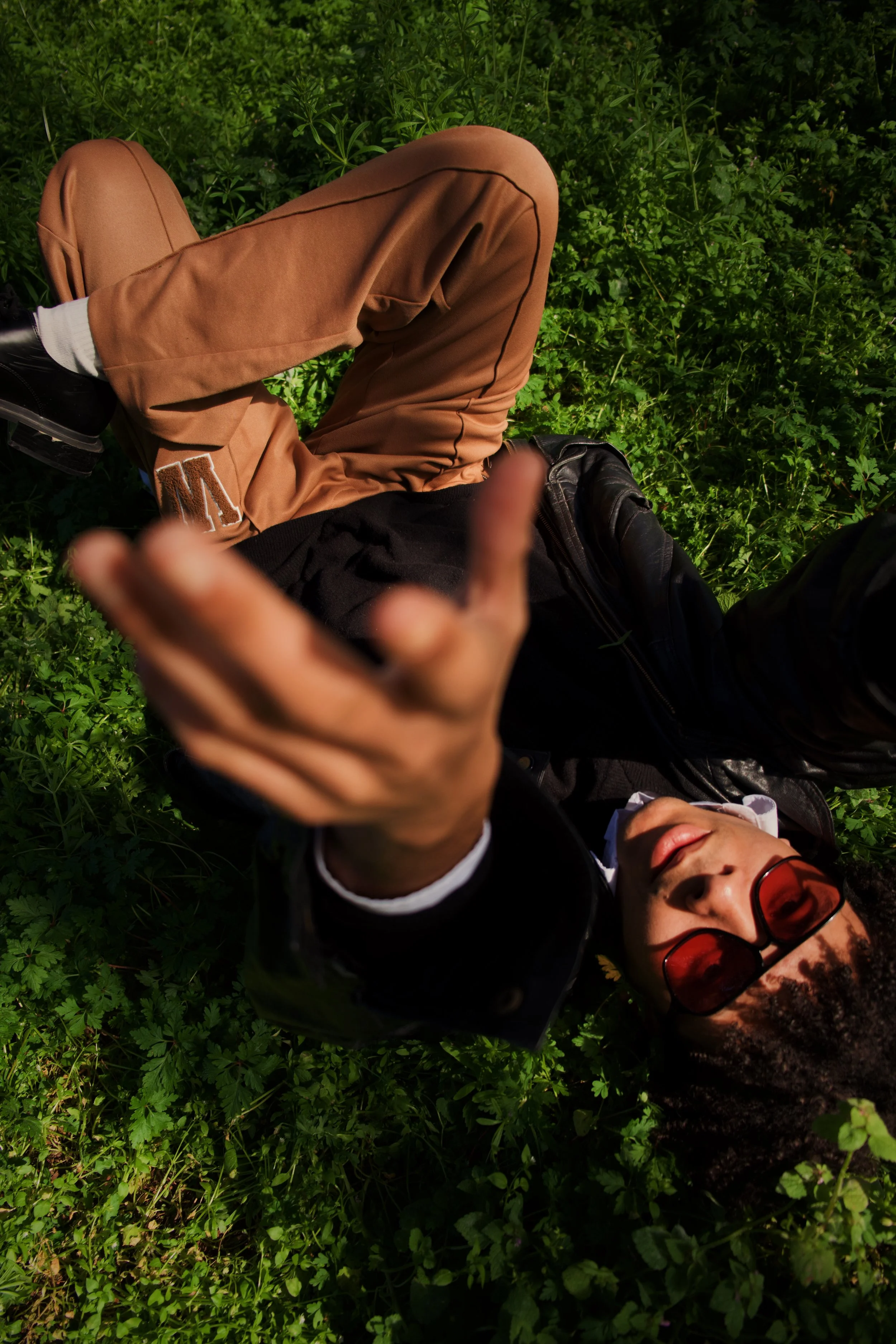 Une personne portant des lunettes de soleil rouges, une veste en cuir noire et un haut blanc, est allongée sur de l'herbe verte et des petites plantes, prenant une photo selfie. Elle porte un bandeau ou un foulard avec un motif. Son visage est détendu et elle semble profiter du moment.
