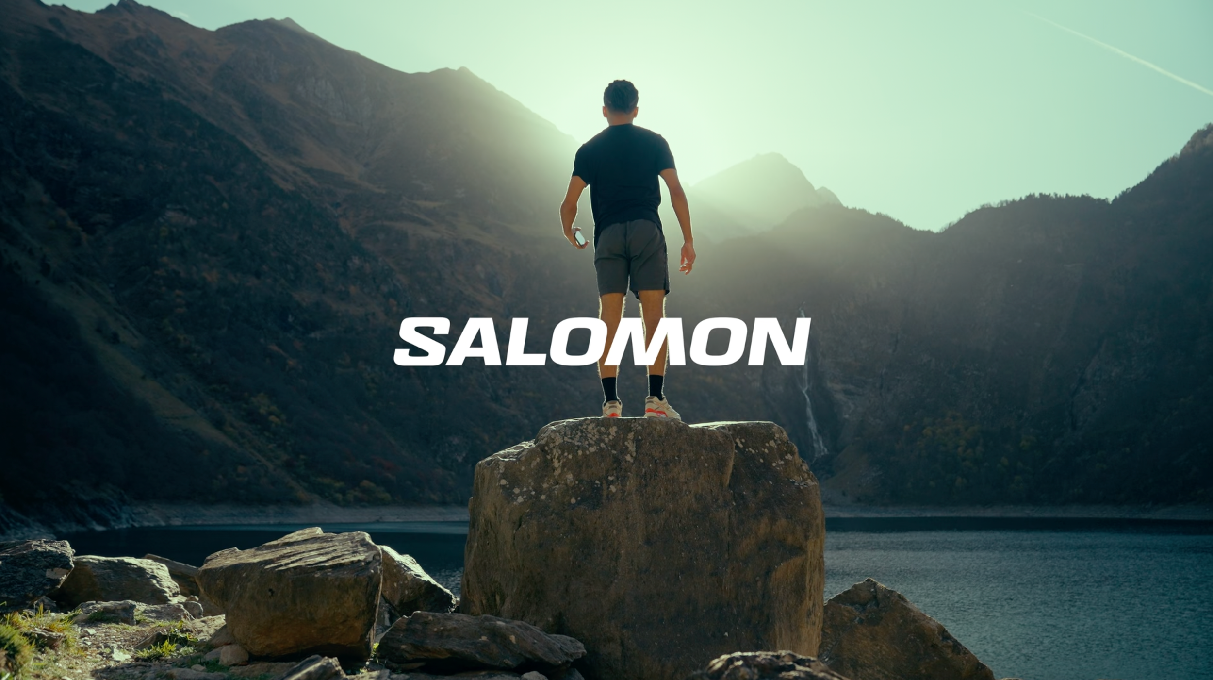 Un homme debout sur une roche au bord de l'eau dans un paysage de montagne, avec le soleil levant ou couchant à l'horizon et le mot « SALOMON » en lettres blanches en bas de l'image.