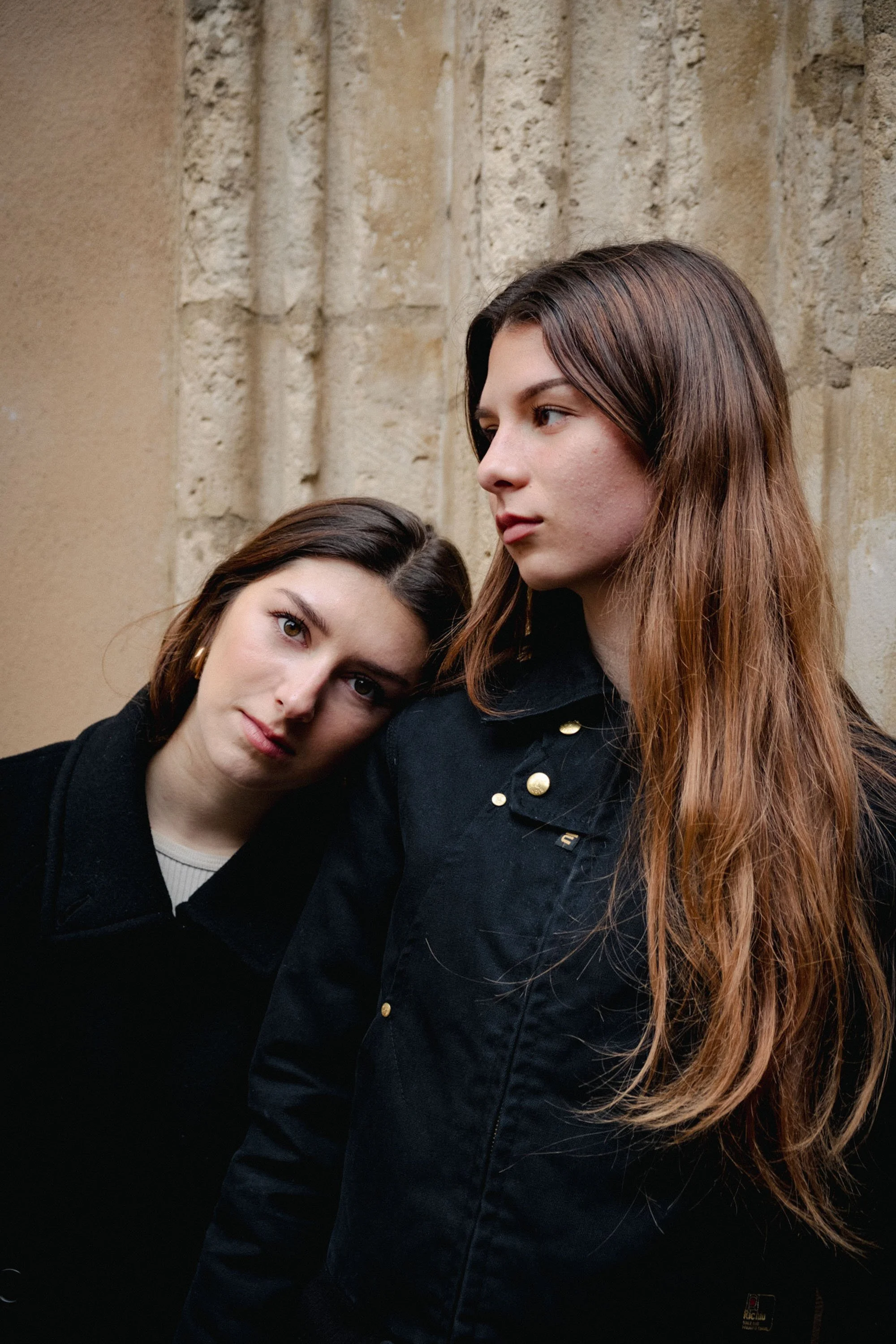 Deux jeunes femmes posent contre un mur en pierre, l'une en regardant vers la droite et l'autre posant la tête sur l'épaule de l'autre, portant des vêtements noirs.