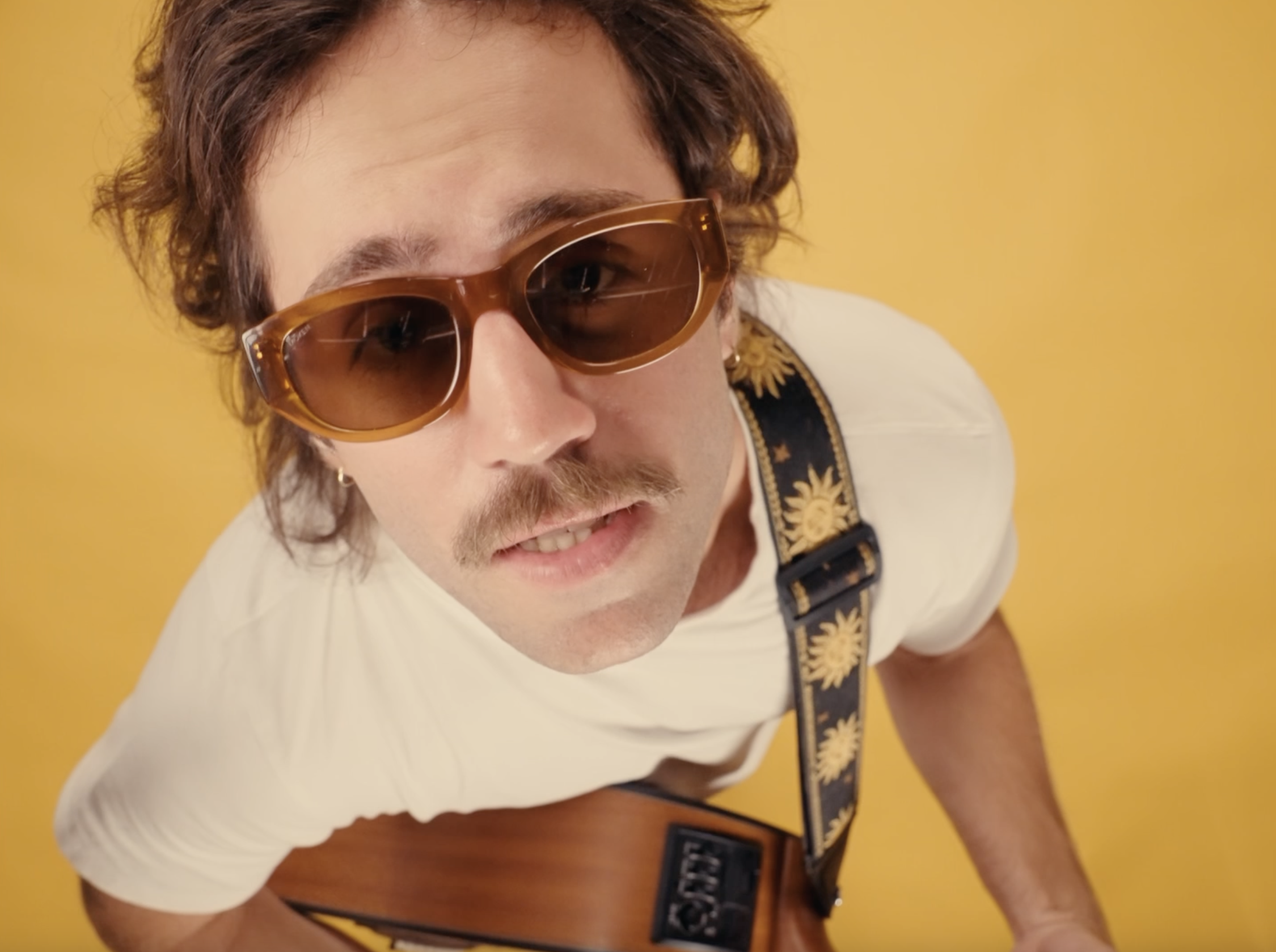 Jeune homme portant des lunettes de soleil marron clair, un t-shirt blanc et une sacoche noire avec une chaîne jaune, posant devant un mur jaune.