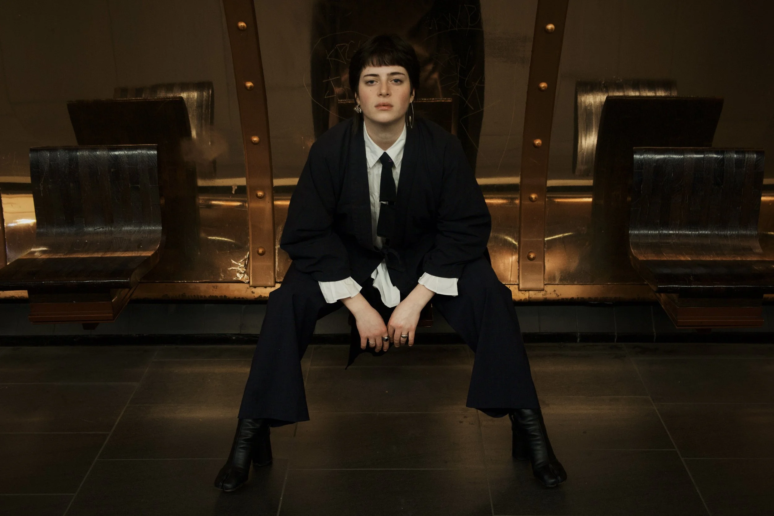 Une femme assise sur un banc en métal dans un espace sombre, portant un costume noir, une chemise blanche et des bottes à talons. Elle regarde droit devant avec une expression neutre.