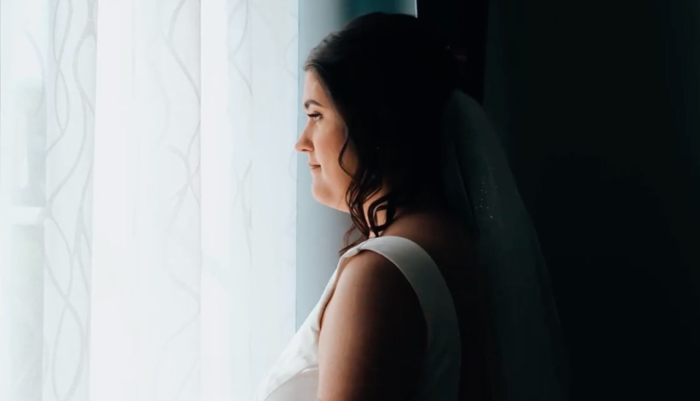 Une femme brune aux cheveux bouclés regarde par une fenêtre avec des rideaux transparents, sa tête légèrement inclinée vers l'avant, dans une pièce sombre.