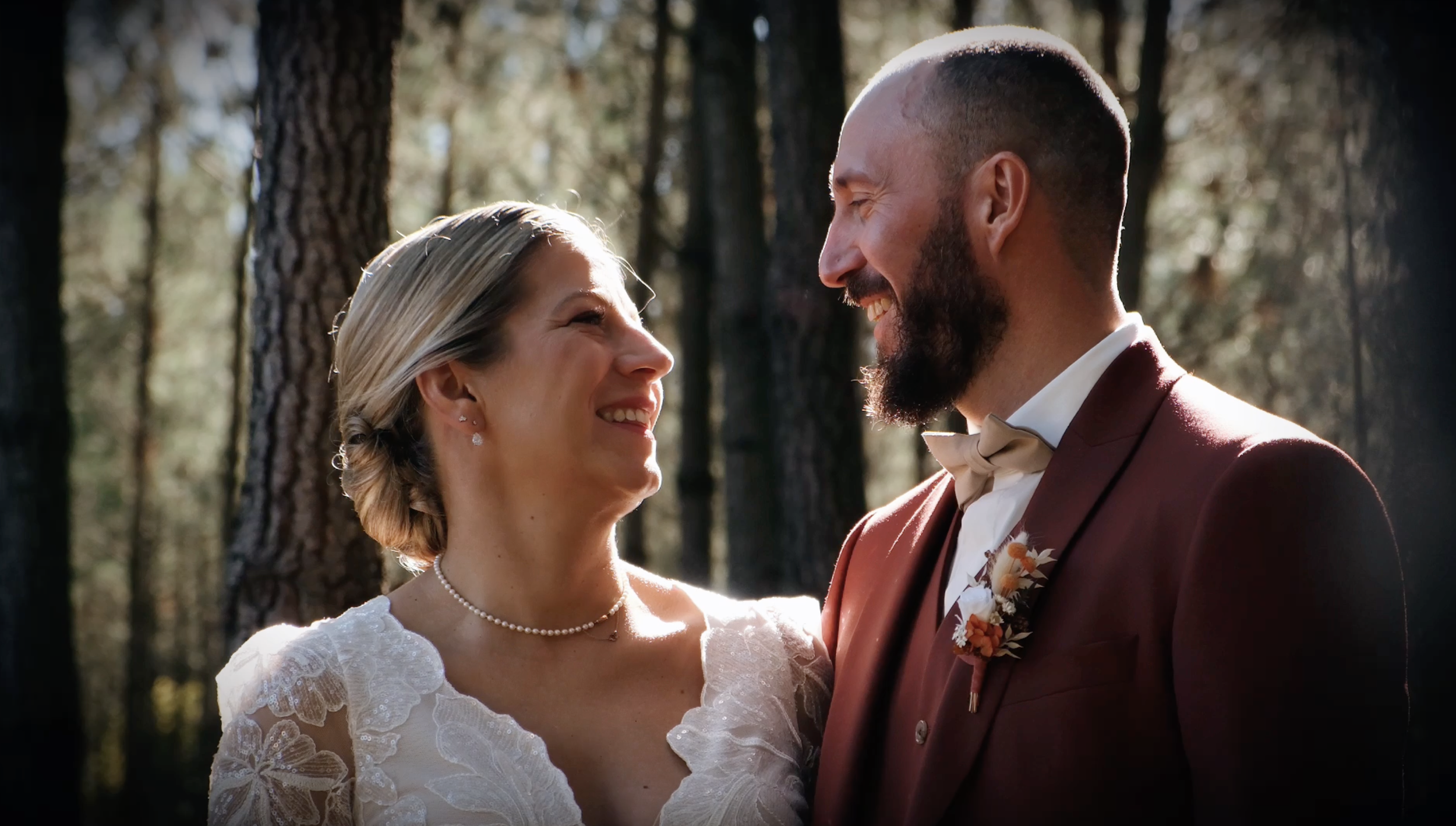 Un couple de mariés regarde dans les yeux, souriant, dans une forêt.