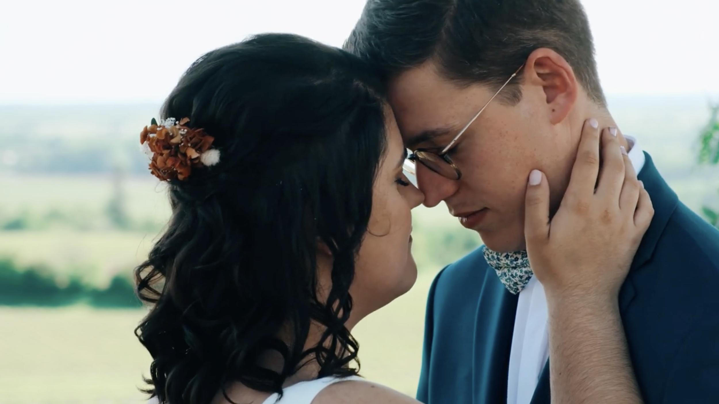 Un couple, une femme aux cheveux noirs avec une coiffure florale et un homme avec des lunettes, se touchent le front et sourient avec tendresse.