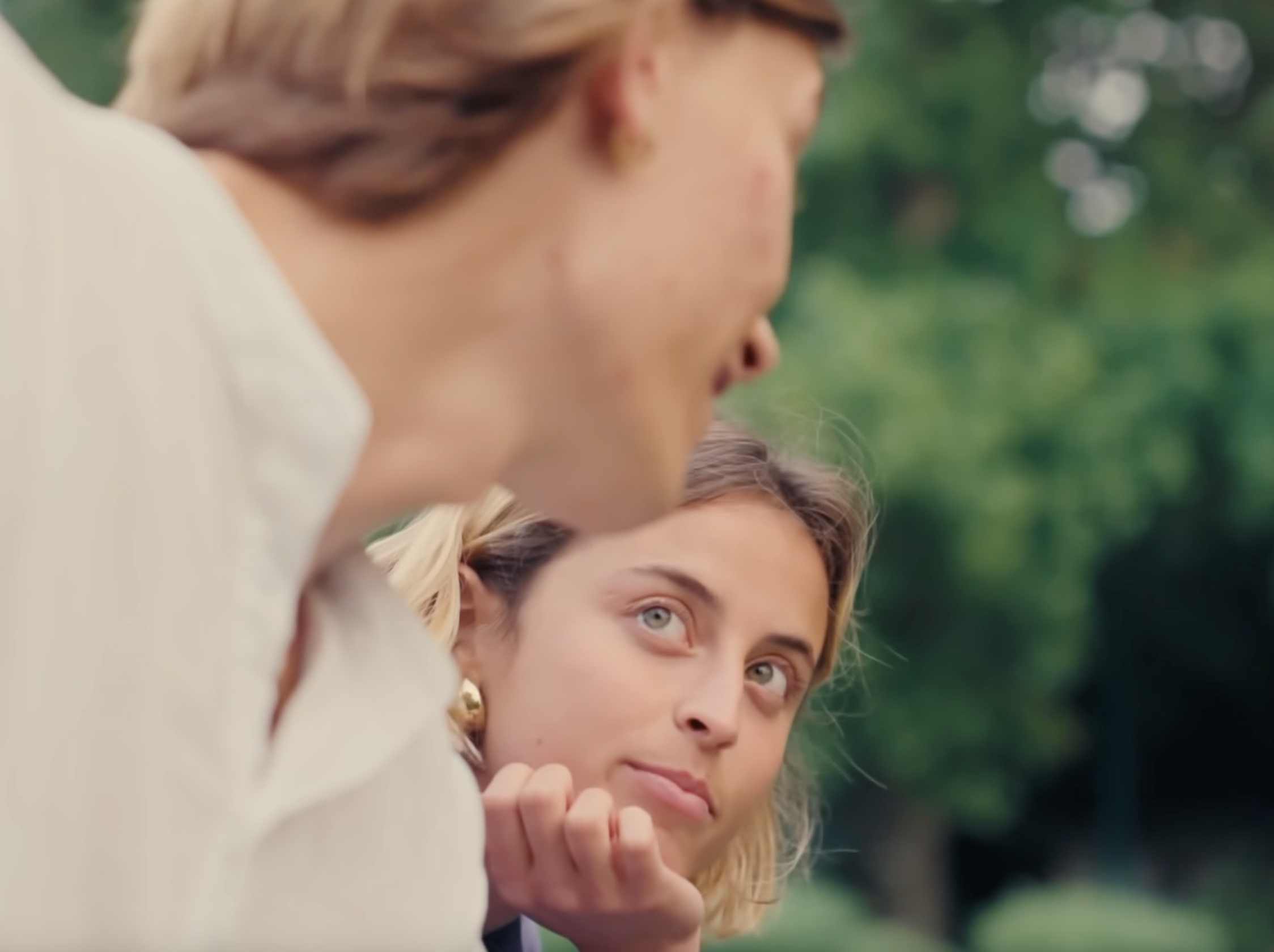 Regard d'une femme blonde avec cheveux ondulés, portant des boucles d'oreilles dorées, regardant vers la droite dans un environnement extérieur verdoyant.