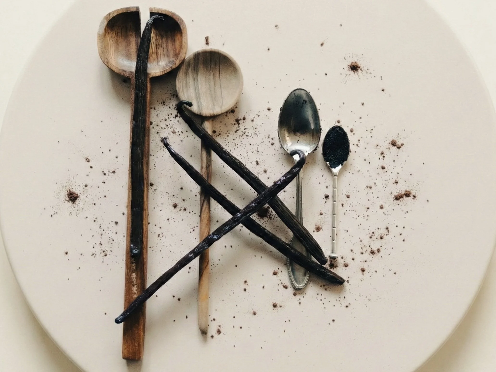 Une assiette blanche avec des cuillères en bois et en métal, quelques grains de cacao et de la poudre de cacao éparpillés dessus.