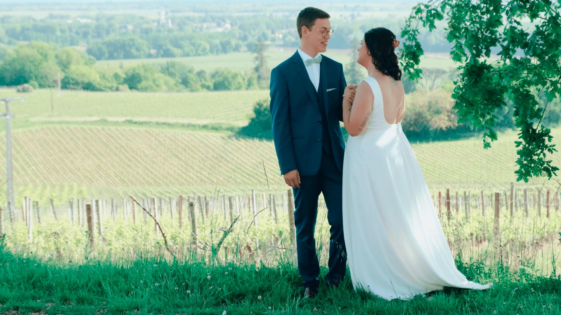 Un couple en tenue de mariage, homme en costume bleu et femme en robe blanche, échange un regard dans un champ avec un paysage de vignobles en arrière-plan, sous un arbre.