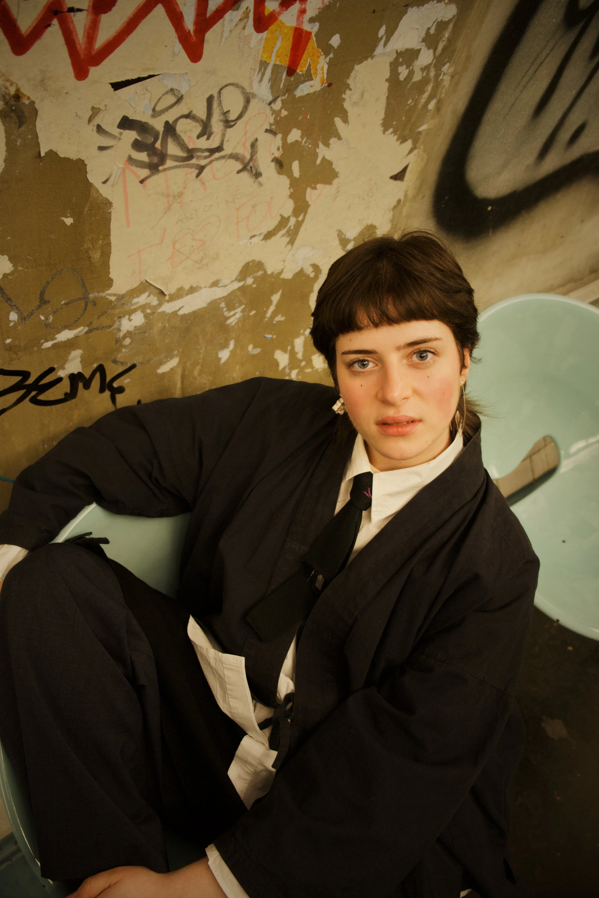 Une jeune femme avec des cheveux courts et bouclés, portant un costume noir, une chemise blanche et une cravate noire, assise dans un endroit urbain avec un mur dégradé en arrière-plan recouvert de graffitis.