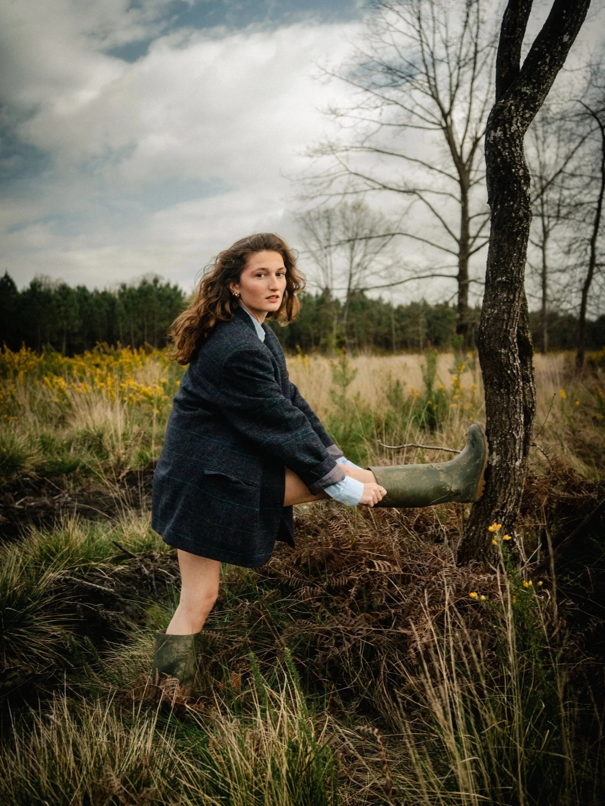 Une femme avec des bottes en caoutchouc, une veste en laine et une chemise en jean, assise dans une prairie avec des fleurs jaunes, tenant une botte en caoutchouc contre un arbre, sous un ciel nuageux, en arrière-plan des arbres dénudés.