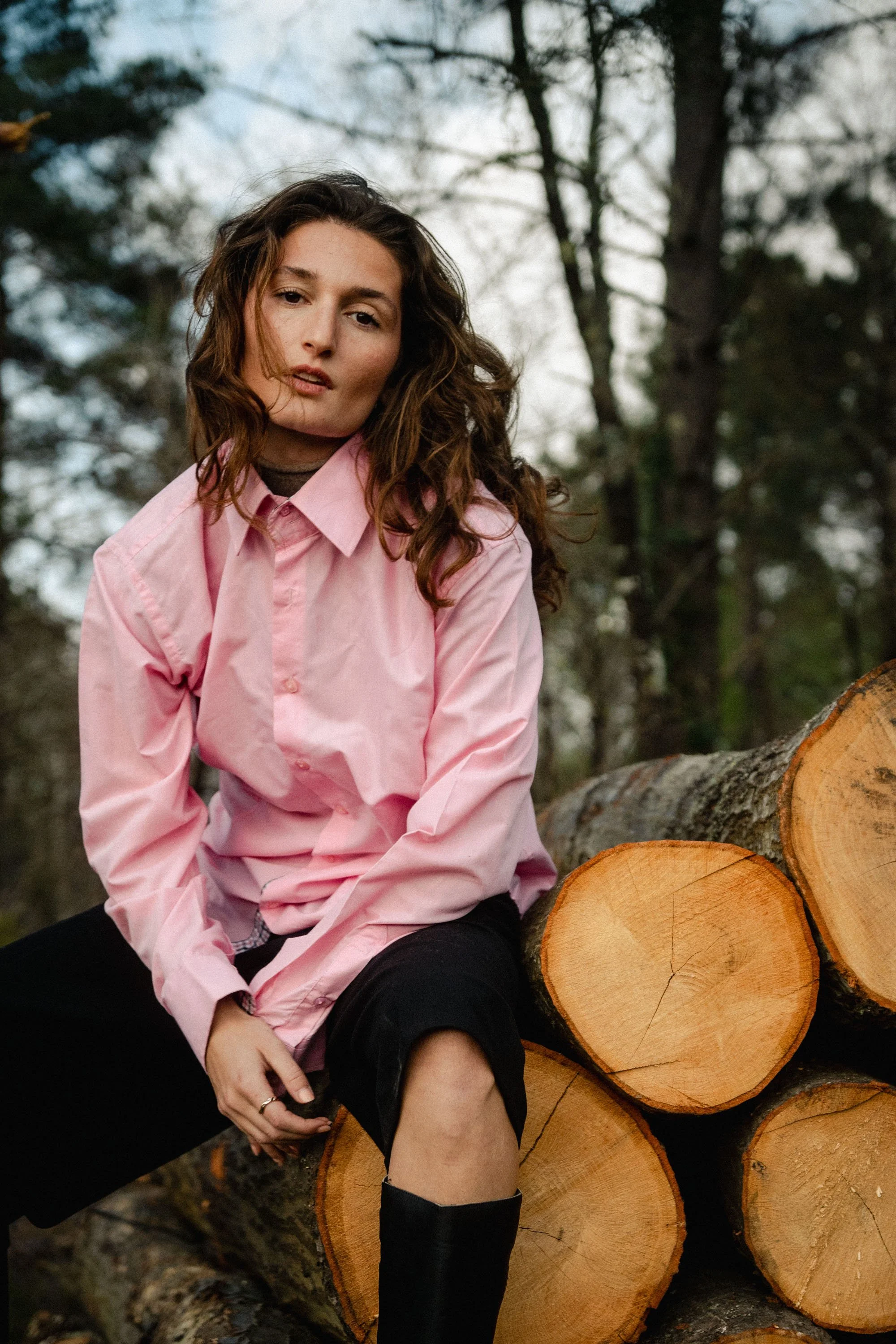 Une femme assise sur un tas de troncs d’arbres dans une forêt, portant une chemise rose et des chaussettes noires, avec une expression neutre.