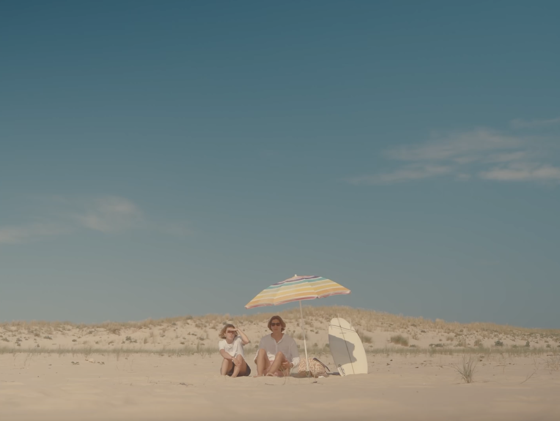 Trois personnes assises sous un parasol à la plage, avec un cerf-volant rose dans le sable, face à une mer calme et un ciel bleu clair.
