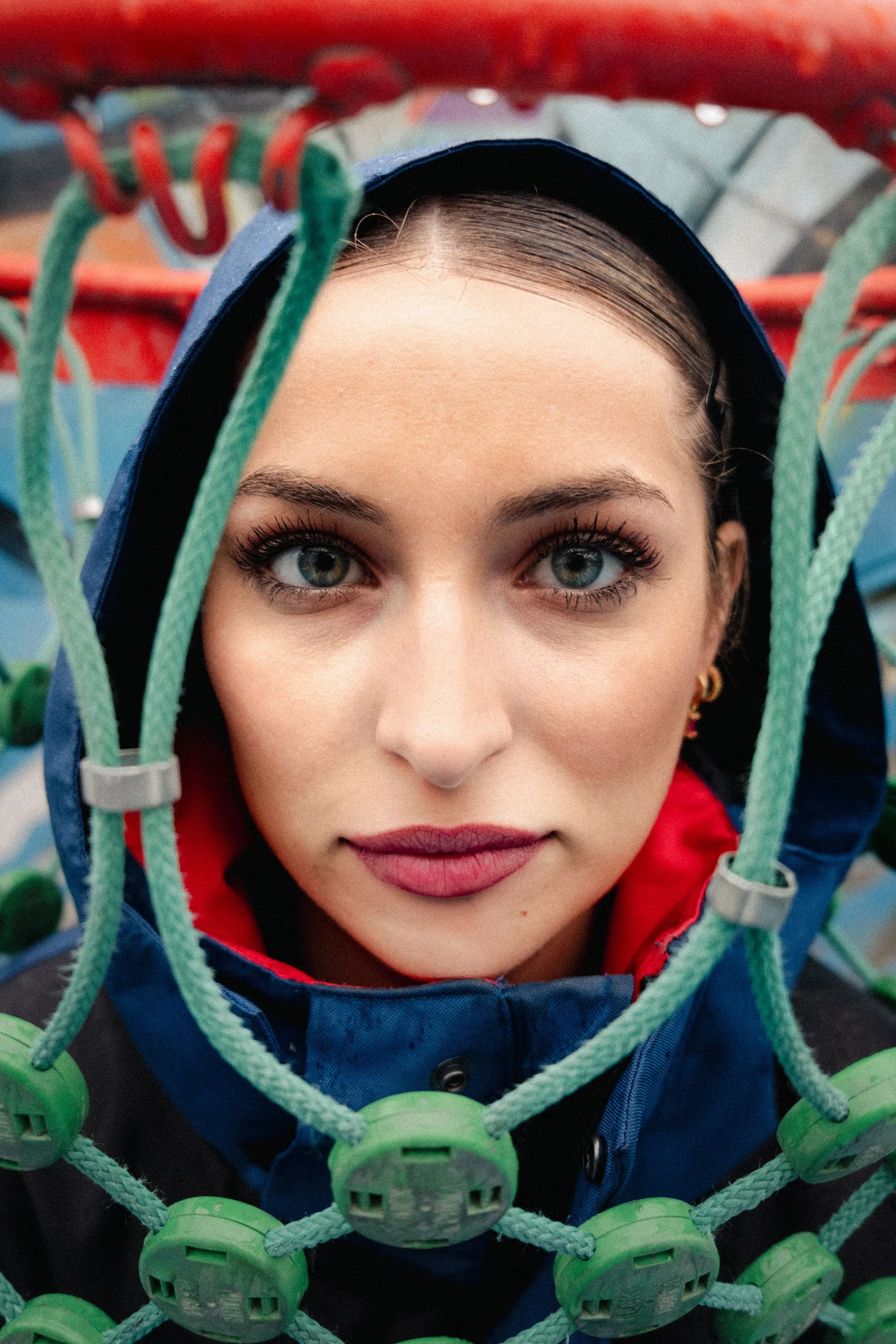 Jeune femme regardant au-dessus, vue de face, à travers une chaîne de sport de couleur verte, portant une capuche bleue et un vêtement rouge, avec des yeux bleus et un maquillage subtil.