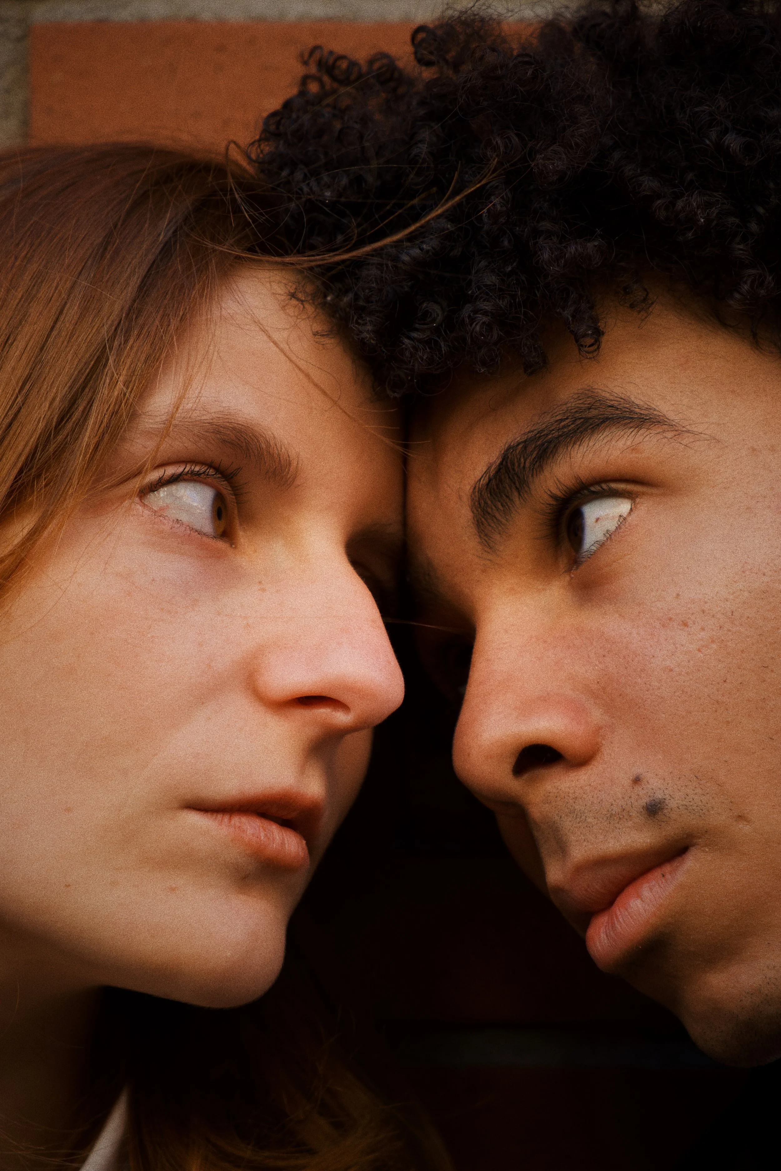Deux personnes, une femme avec les cheveux roux et une homme avec des cheveux bouclés noirs, regardent intensément l'un l'autre, leurs visages très proches.