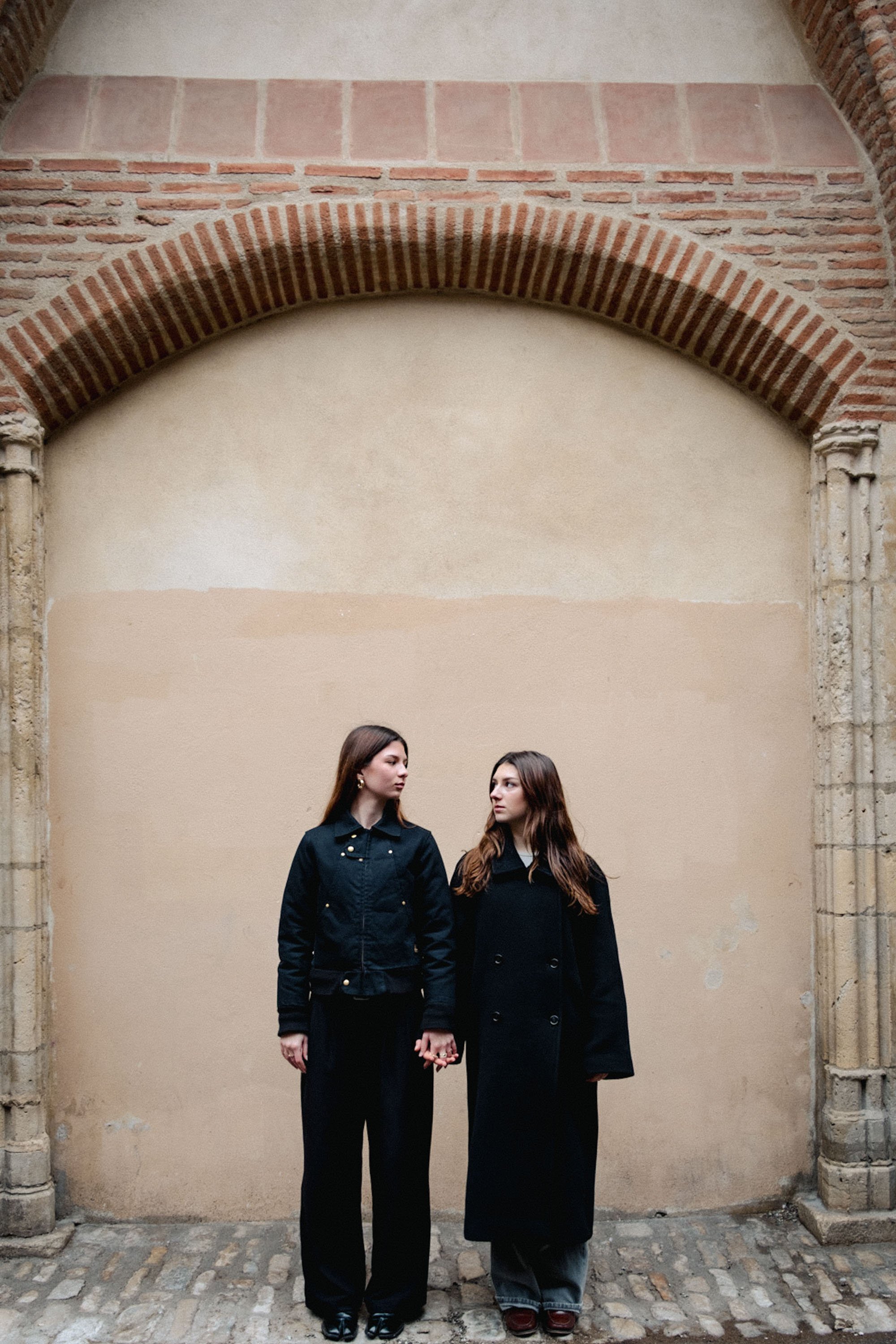 Deux jeunes femmes debout côte à côte, portant des vêtements sombres, sous une grande arche en briques dans un environnement ancien.