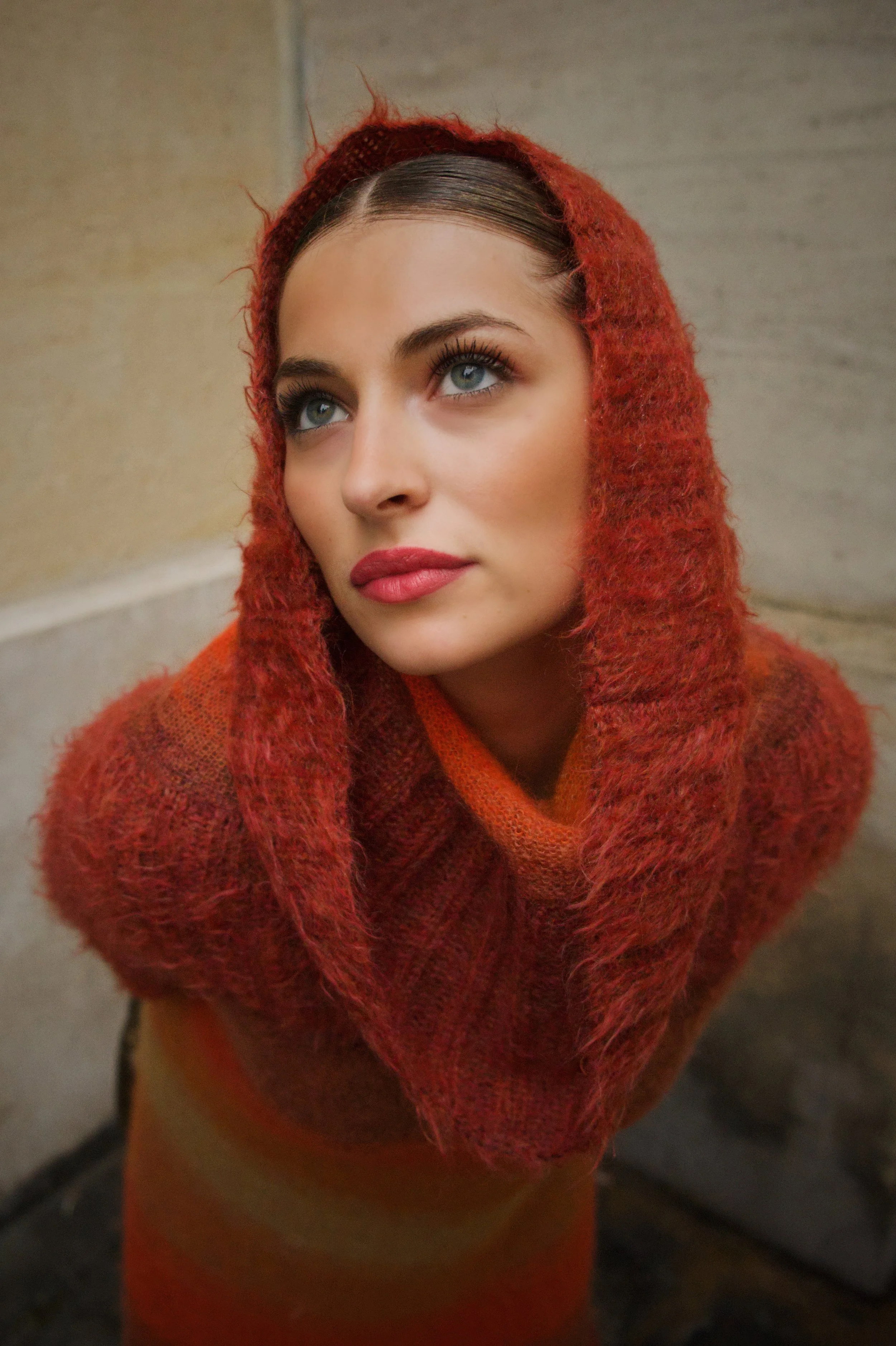 Jeune femme portant une écharpe et un pull rouges ou orange, avec un maquillage sophistiqué, regardant vers le haut.