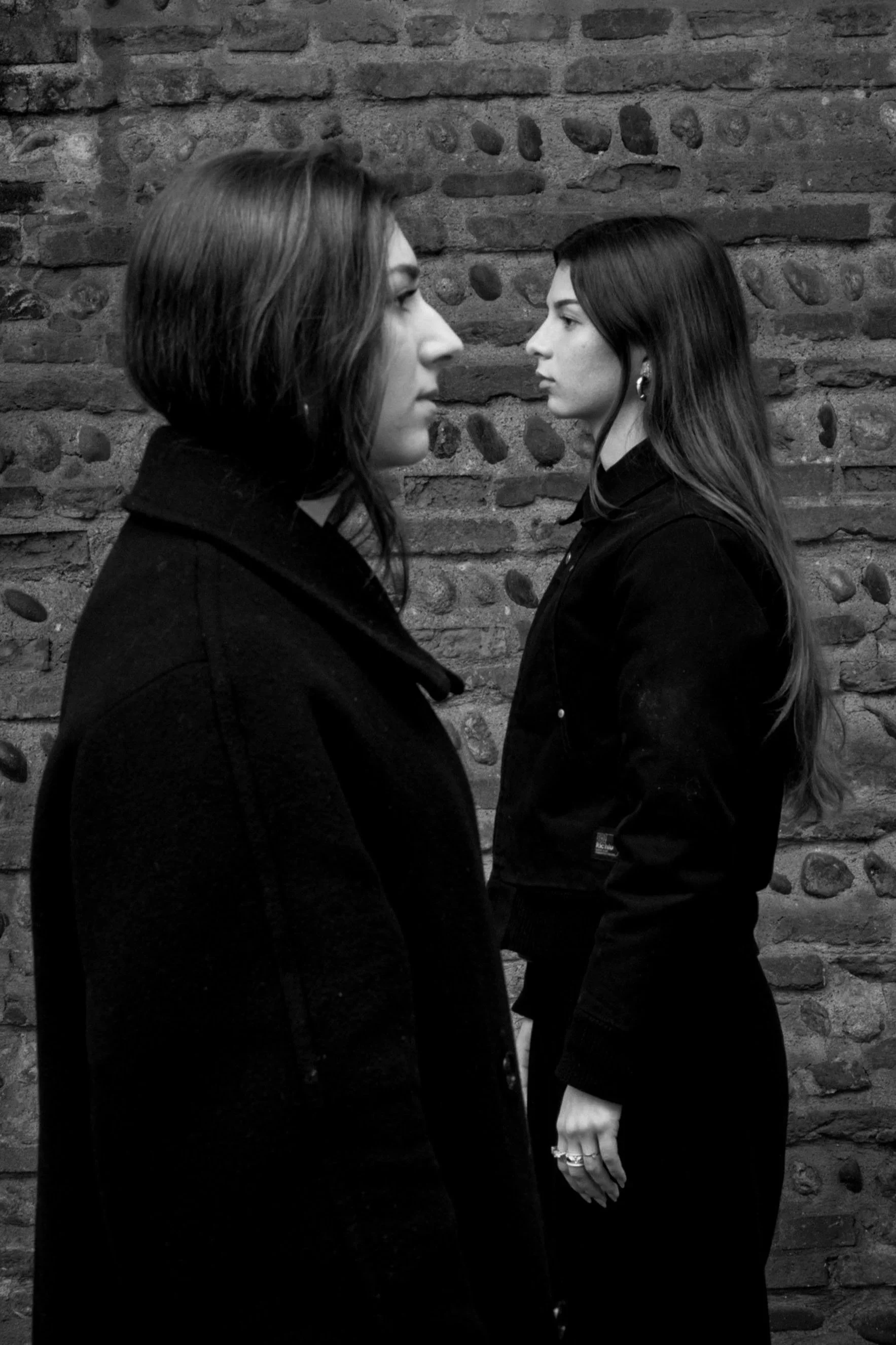 Deux femmes se font face, vues de profil, devant un mur en pierre, avec un style en noir et blanc.