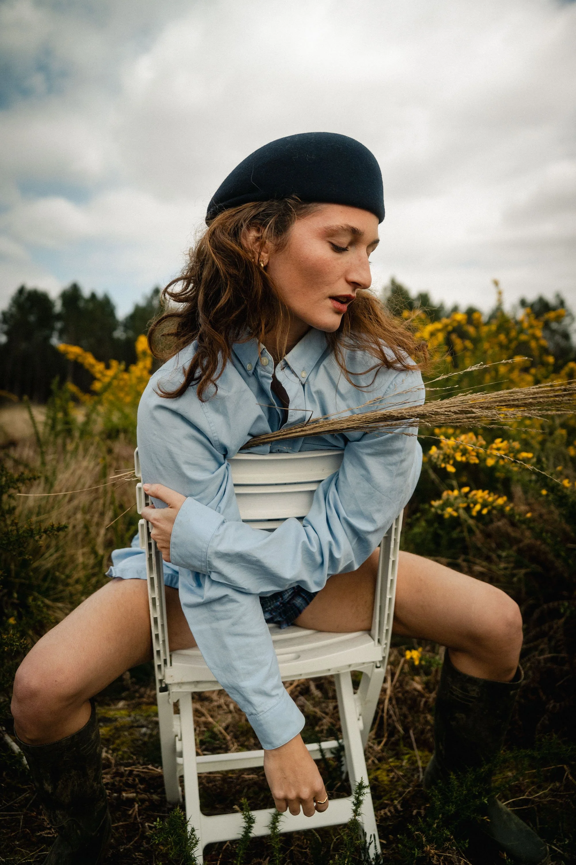Une jeune femme aux cheveux bouclés et portant un bonnet noir, assise sur une chaise en plein air. Elle porte une chemise bleue et une jupe courte, avec des bottes noires. Elle tient une herbe dans la main, dans un champ avec des buissons jaunes en arrière-plan, par une journée nuageuse.