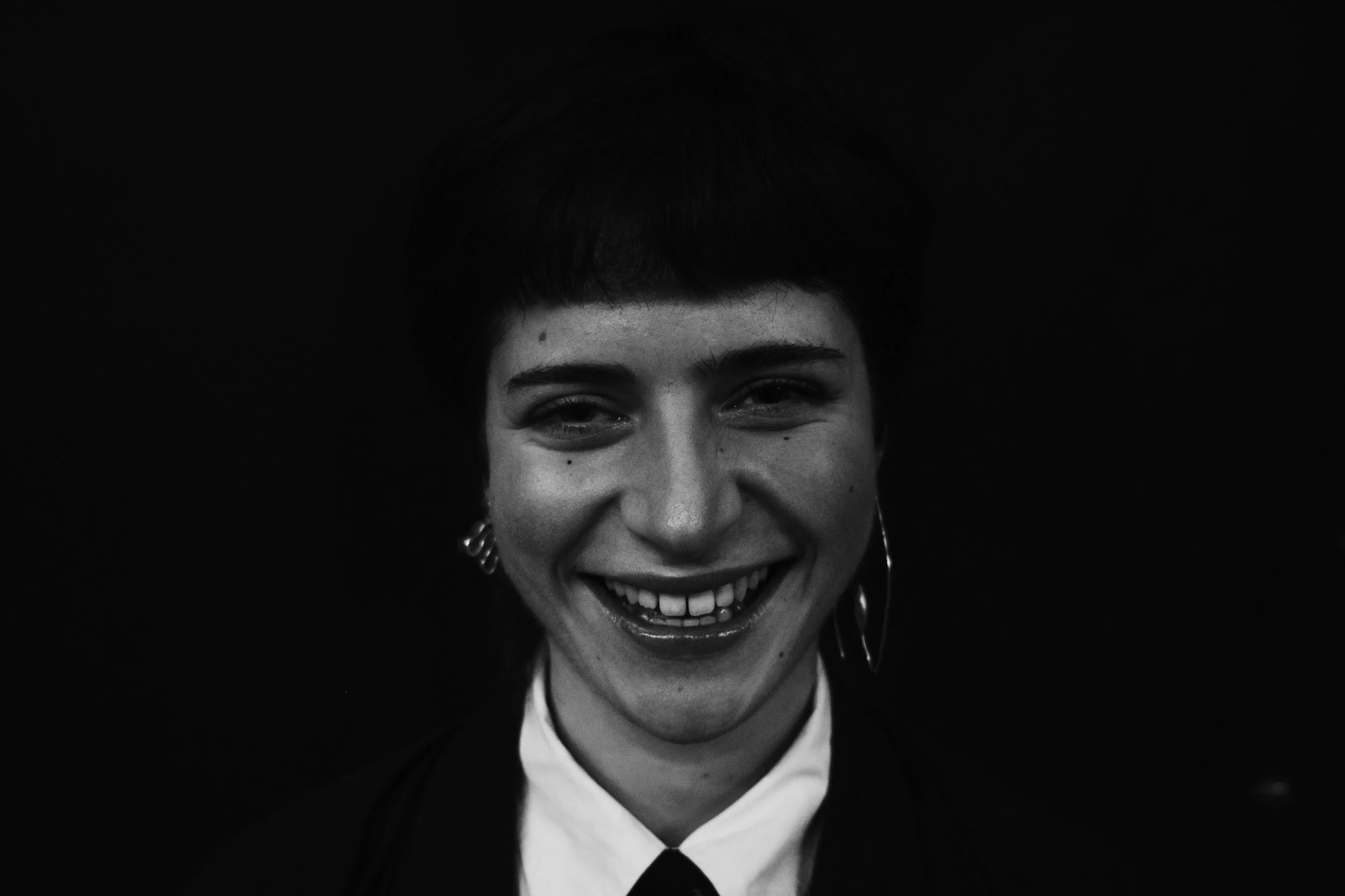 Portrait en noir et blanc d'une jeune femme souriante, portant des boucles d'oreilles et une veste noir avec un chemisier blanc, fond sombre.