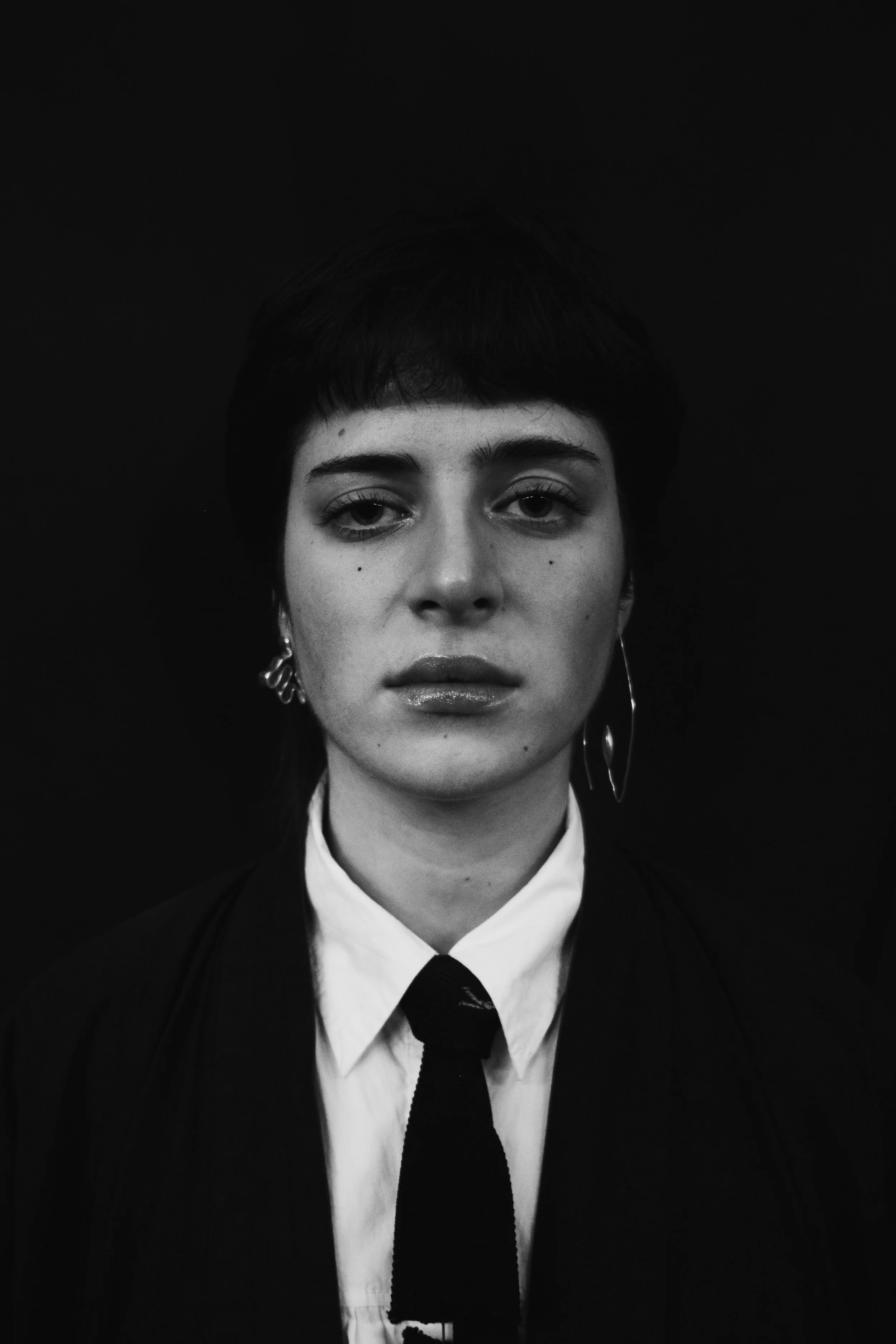 Portrait en noir et blanc d'une femme avec une coupe courte, portant une chemise blanche, un nœud papillon noir et des boucles d'oreilles contrastantes, avec une expression neutre contre un fond sombre.
