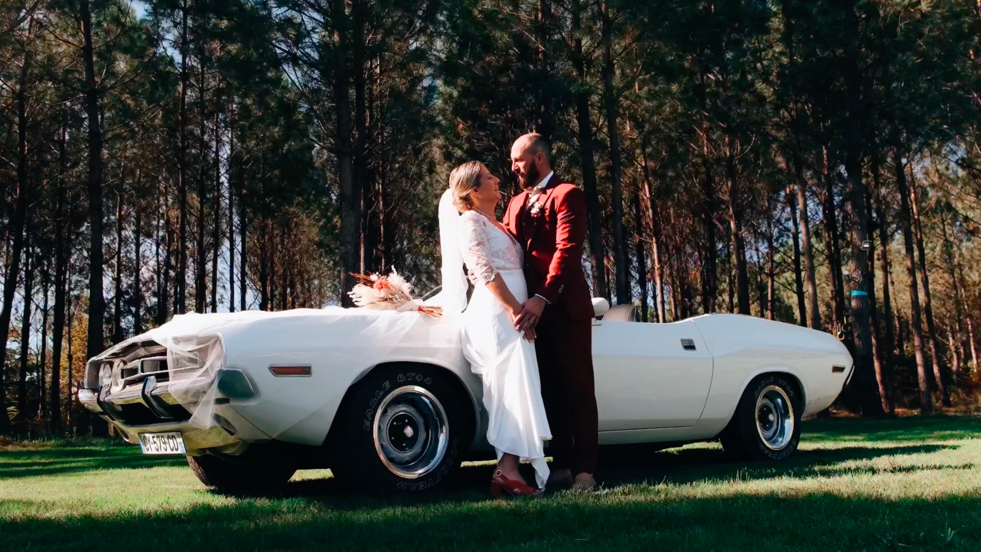 Un couple en mariage, en costume et robe blanche, se tenant la main devant une voiture de collection blanche, dans une forêt.
