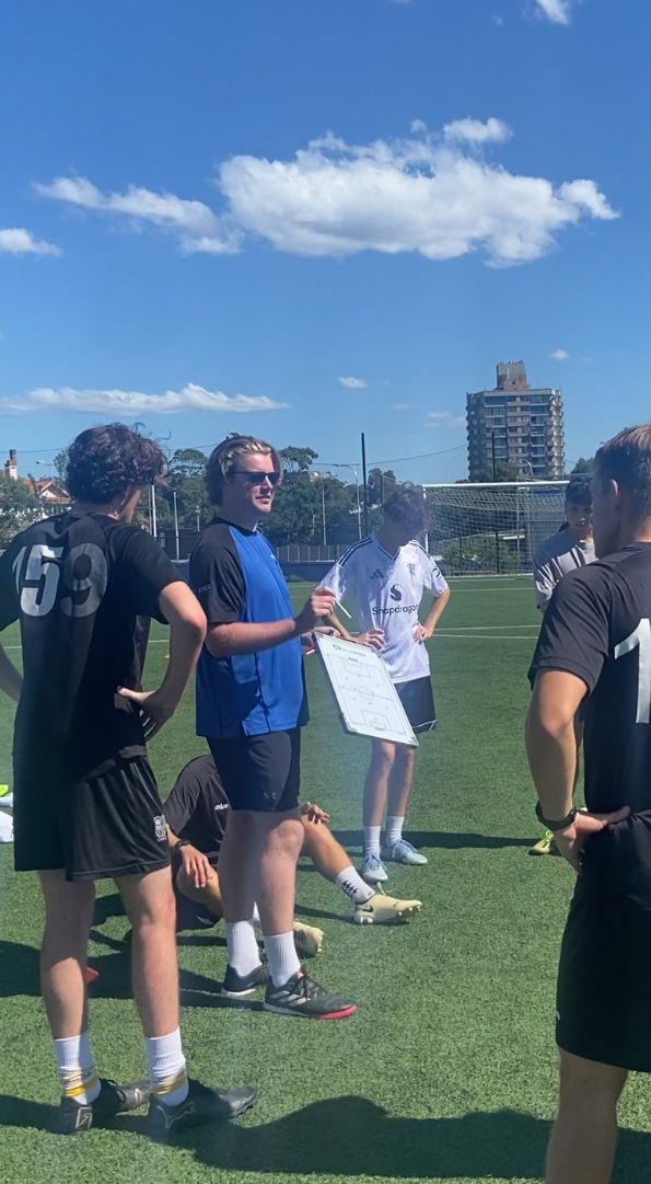 Harbour Football coach giving instructions to a team of young players on a sunny outdoor soccer field, with a cityscape and clouds in the sky in the background.