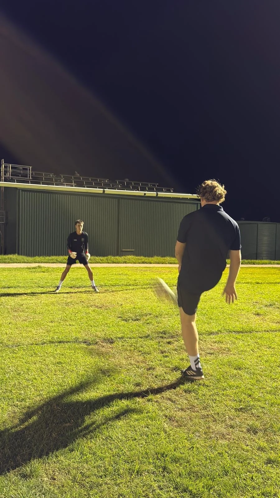 Two young men playing baseball on a grassy field at night, one is pitching while the other is ready to catch, with a dark building in the background.