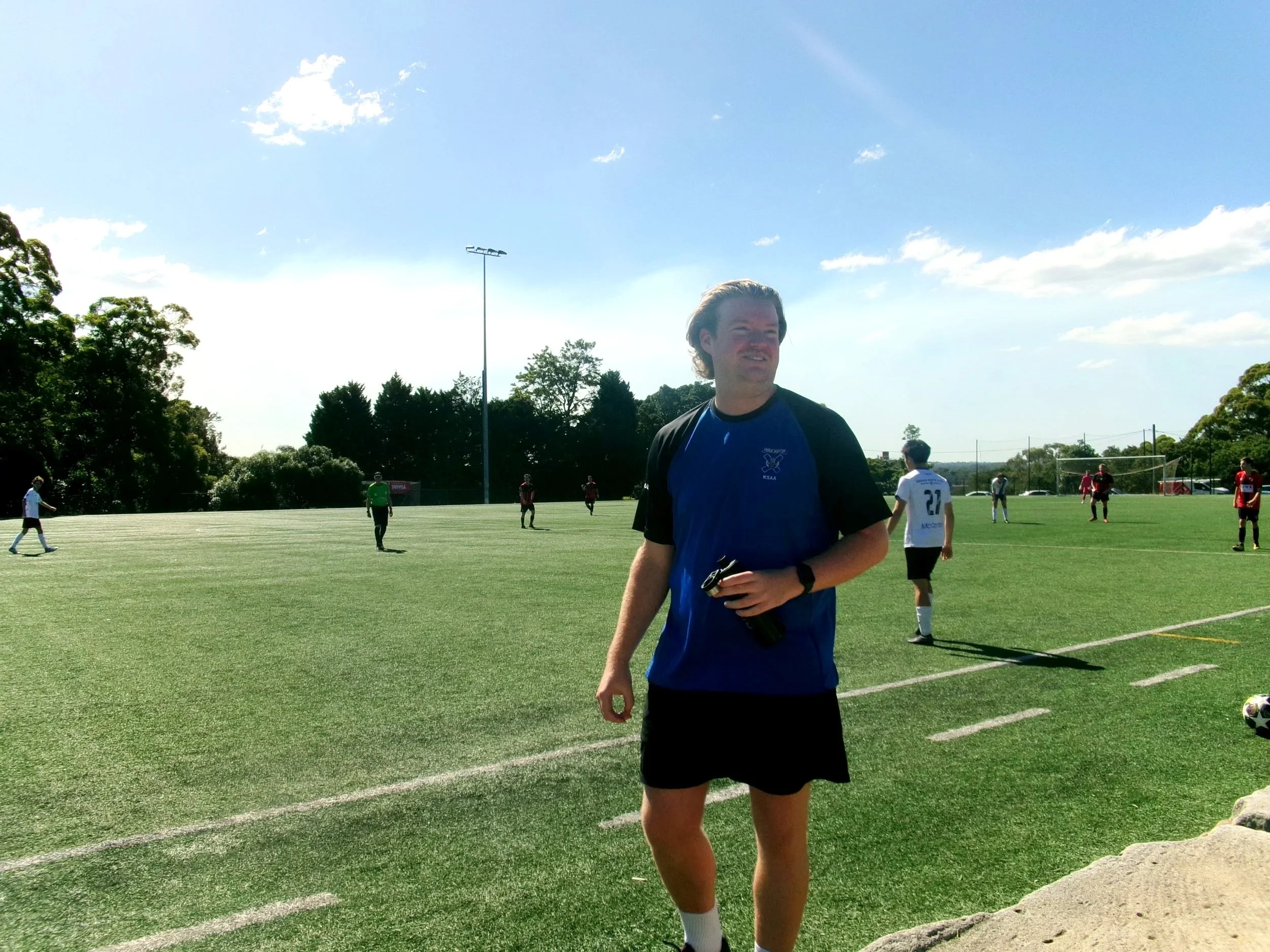 A Harbour Goalkeeper coach in a blue and black sports shirt and black shorts standing on a soccer field, with other players in the background and a partly cloudy sky overhead.