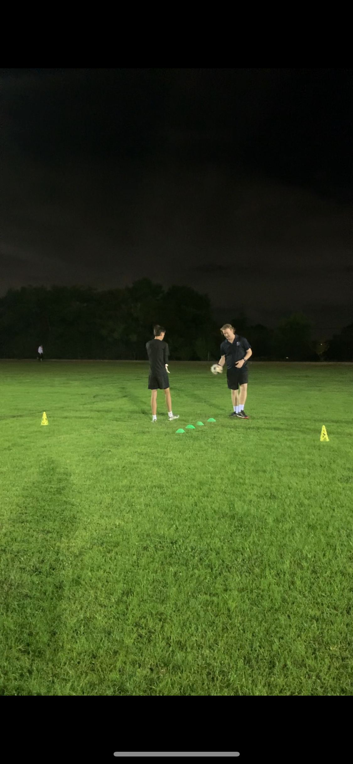 Two young men running goalkeeper training drills on a grassy field at night, with training cones and markers, and night sky in the background.