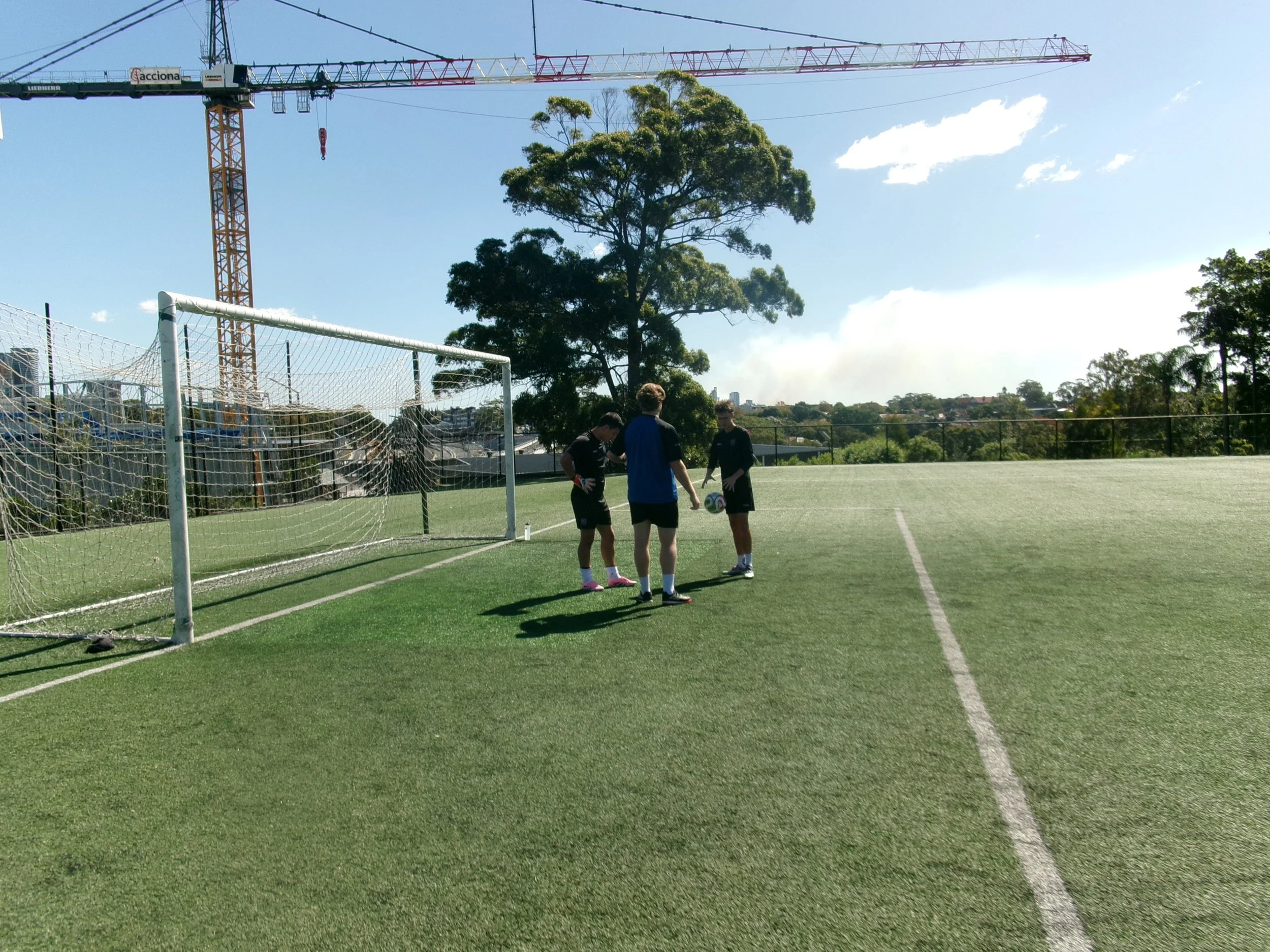 Three goalkeepers on a soccer field preparing to kick a soccer ball with a goalpost nearby. Harbour Goalkeeper coach providing advice and insights on how to imporve during a private small group session