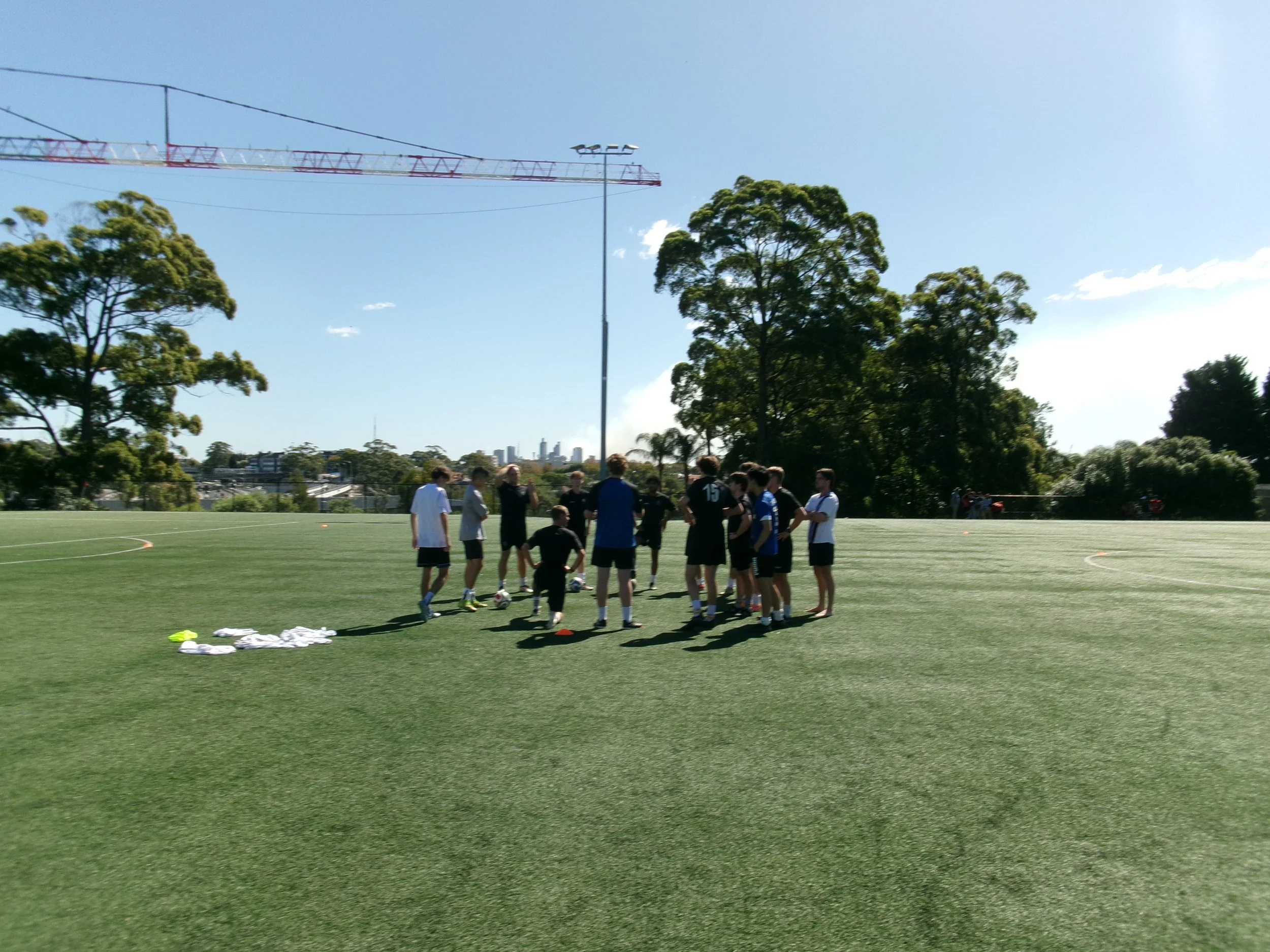 A group of young soccer players gathered on a grassy soccer field with Harbour Goalkeeping coach, with trees and a city skyline in the background during the day. Soccer balls are on the ground, and a pile of white and yellow items is nearby.