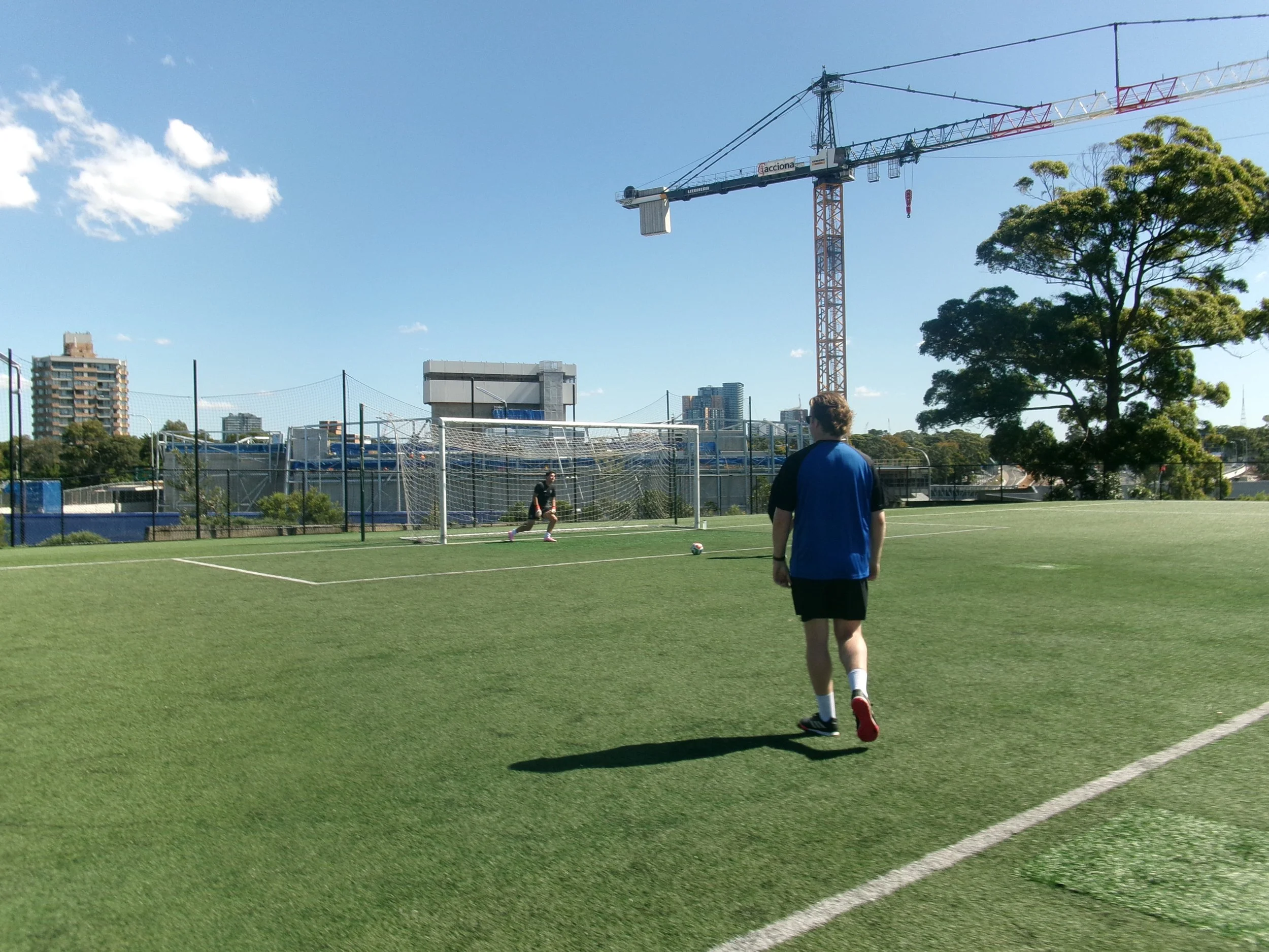 A Harbour Goalkeeping coach in a blue shirt and shorts walking on a soccer field toward a goal with a soccer ball, with a goalkeeper practicing in front of the goal