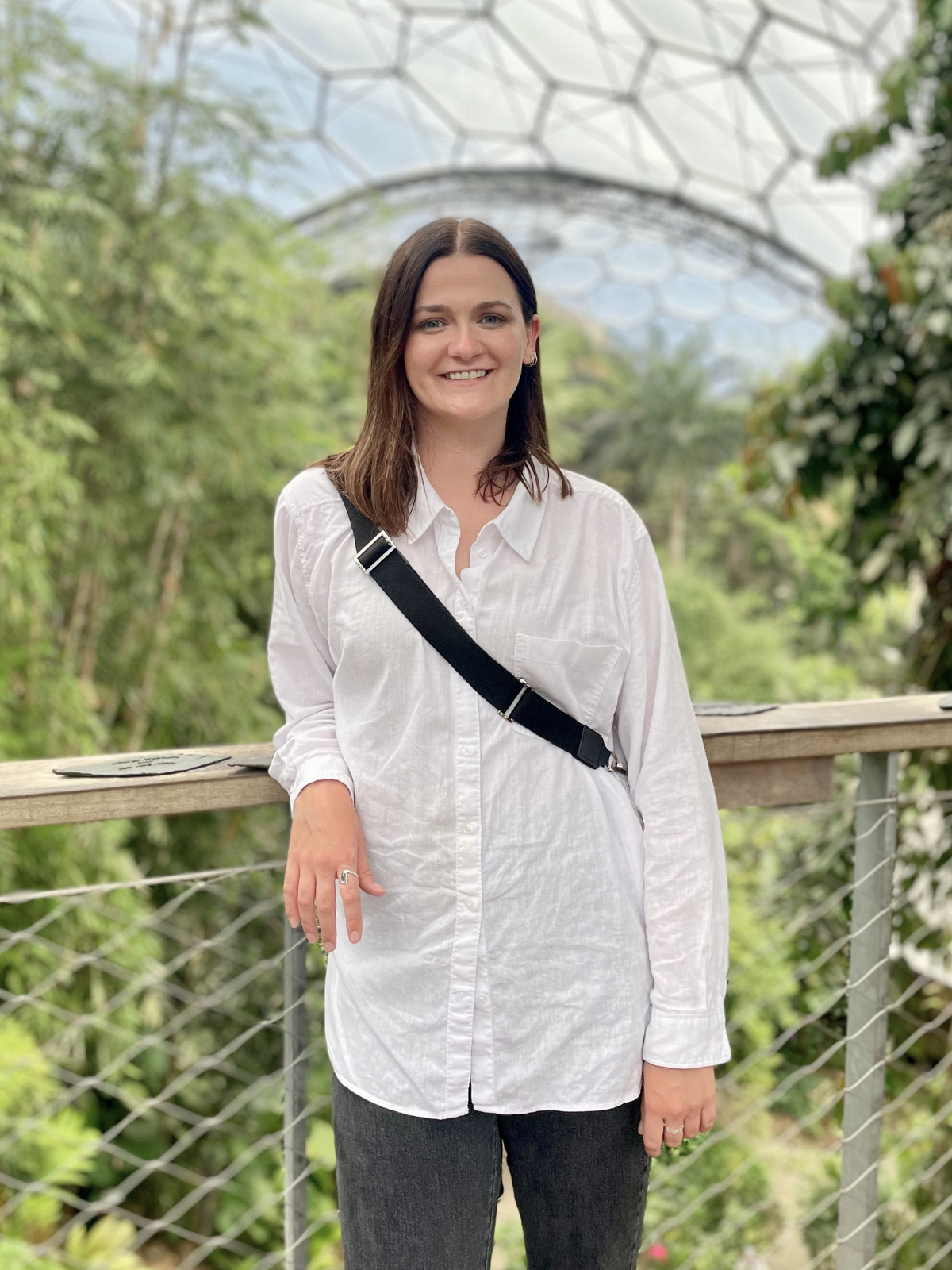 A woman wearing a white shirt and black pants standing on a boardwalk with green trees and a geodesic dome in the background.