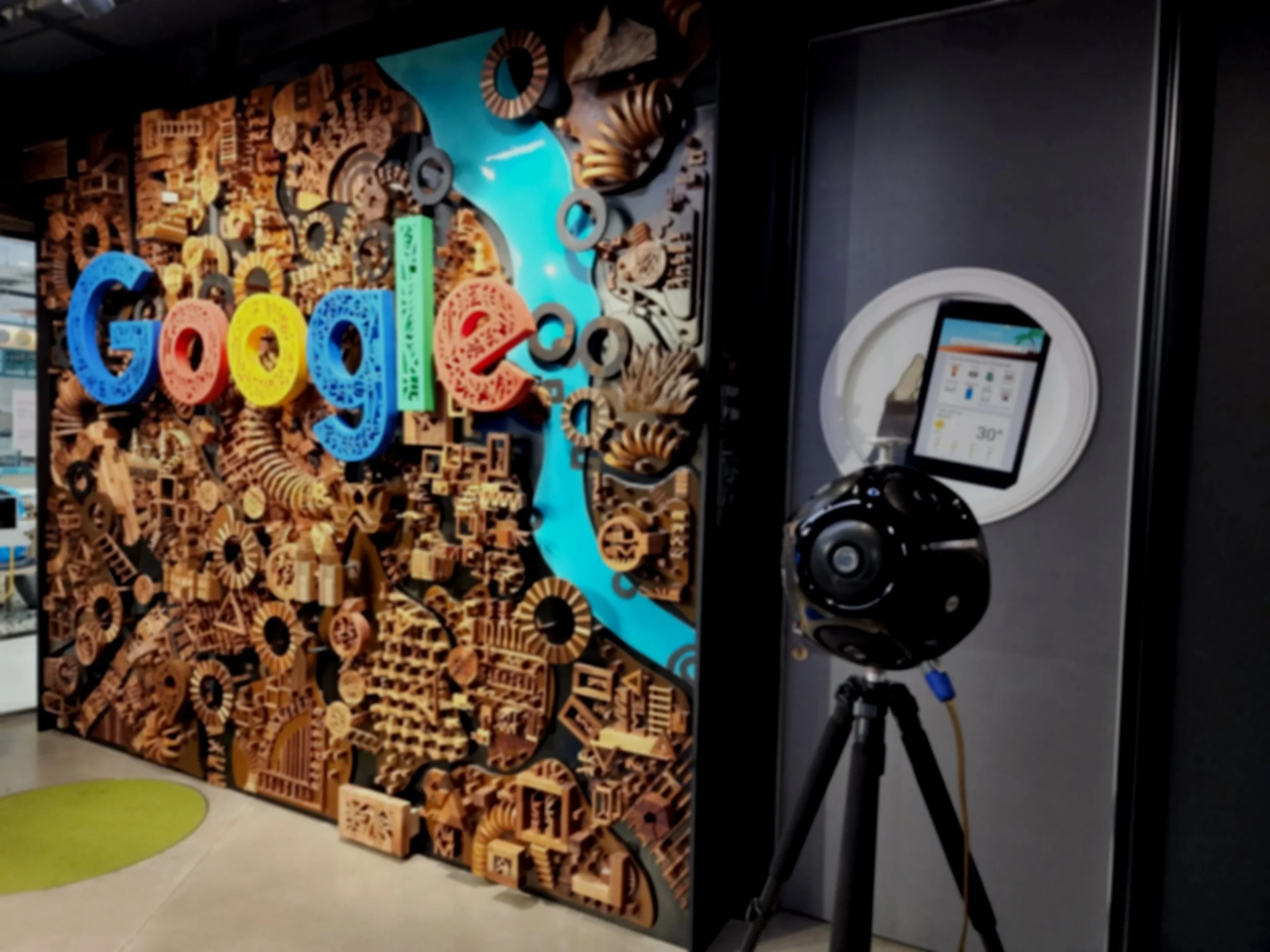 Colorful artwork with the word "Google" in large, multicolored letters, surrounded by wooden puzzle pieces and black circular elements, displayed on a wall with a blue wave design, and accompanied by a digital screen and a tripod in an indoor setting.