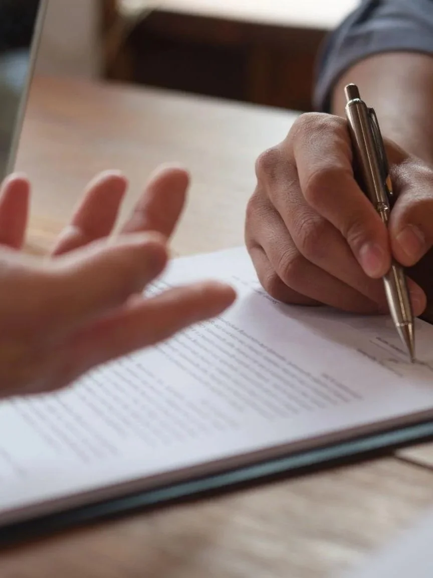 A person is writing on a paper with a silver pen while another person’s hand is reaching out towards the paper.
