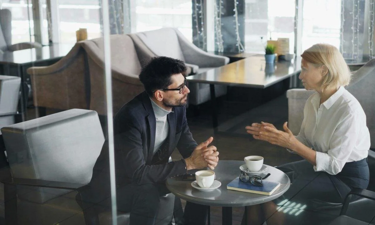 Two people having a conversation at a small table in a cafe, with coffee cups, a notebook, and eyeglasses on the table.