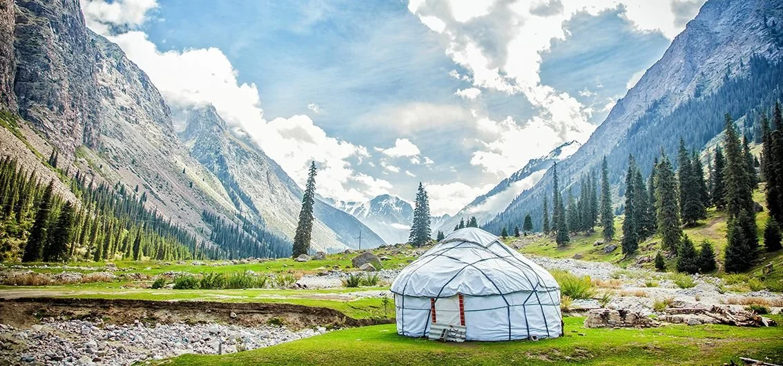 Ein Jurt im Tal mit Bergen und Tannen im Hintergrund, umgeben von Wiesen und steinigen Flüssen, bei sonnigem Himmel.