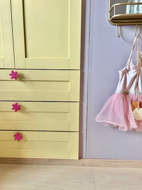 Et barnerom med en gul kommode med tre skuffer, hver med en rosa blomsterknapp som håndtak, og en dress eller kjole med rosa tutu som henger på en knagg på en vegg.