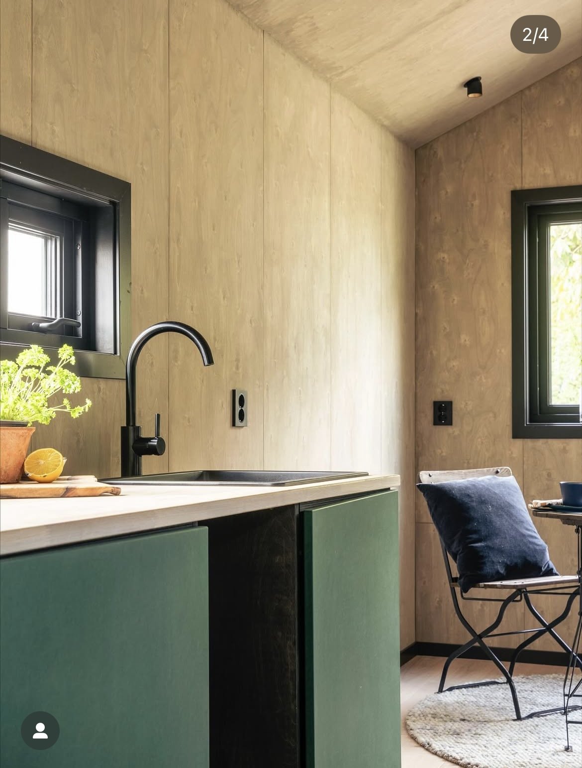 Kjøkken med lyst trebord, svart kran, grønne skuffer, vindu med svart ramme, stue med lenestol og pute, og vindu som slipper inn lys. Foto: Thomas Larsen 