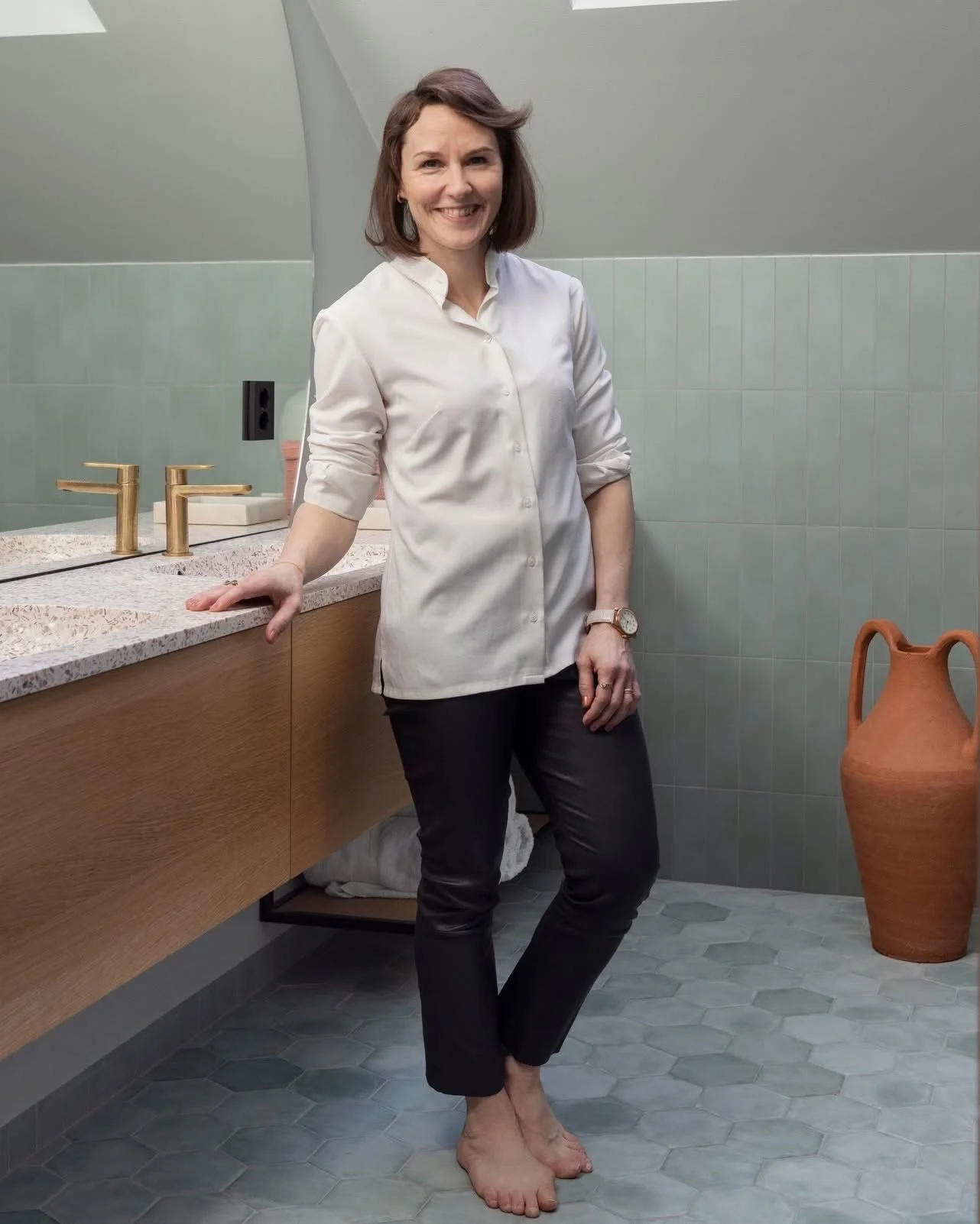 Kvinne står i en badstue, smiler, har på seg hvit skjorte og svarte bukser, med barføtt. Foto: Filippa Tredal