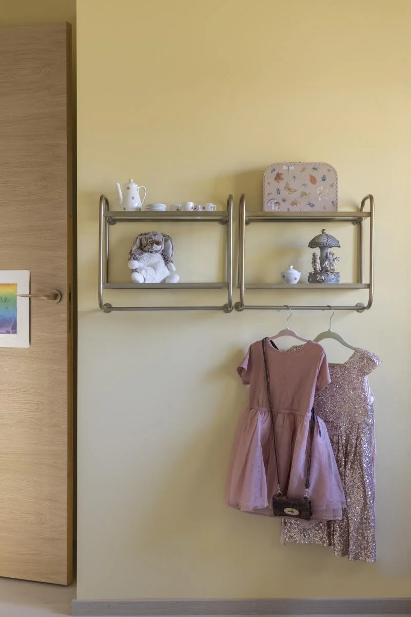 Hyller med keramikk, en bamse, klær som en rosa kjole og en glitterkjole henger på en vegg.